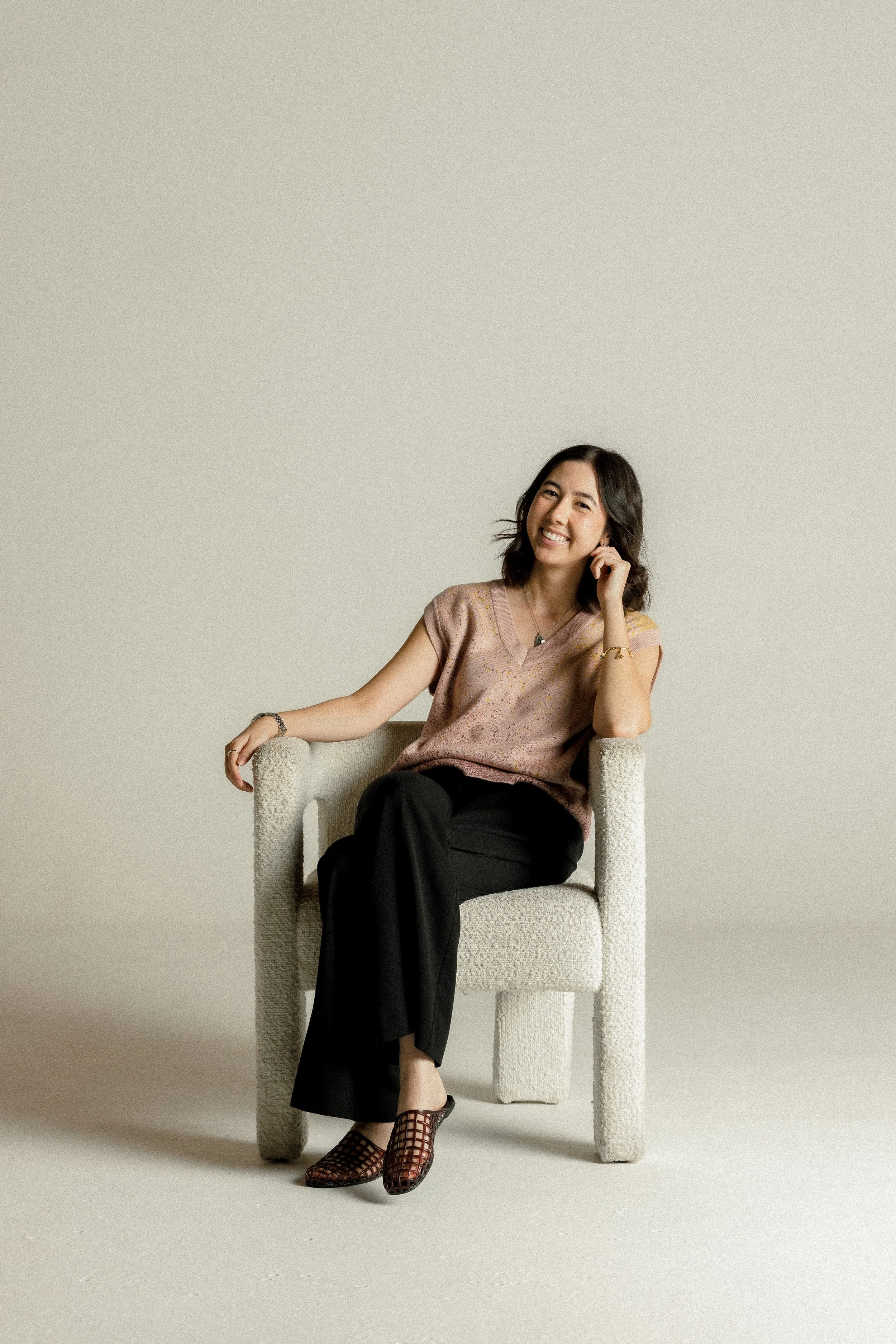 A woman sitting in a beige armchair, smiling, with her head tilted slightly and her hand resting near her face, wearing a short-sleeved, pinkish-beige top, black pants, and patterned open-toe shoes, against a plain, light gray background.