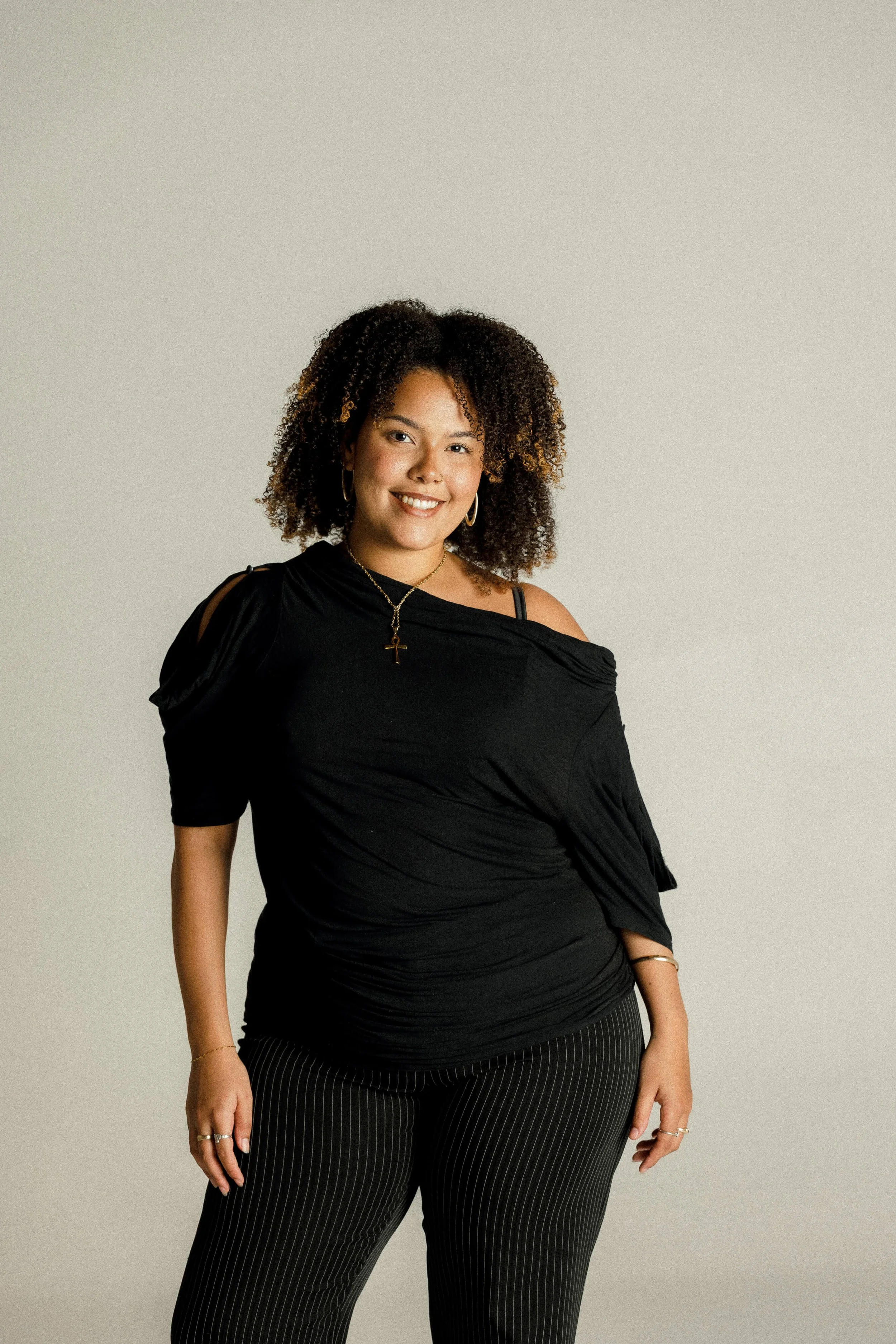 Elsie smiling and wearing a black top with cutout shoulders, gold jewelry, and black pinstriped pants.