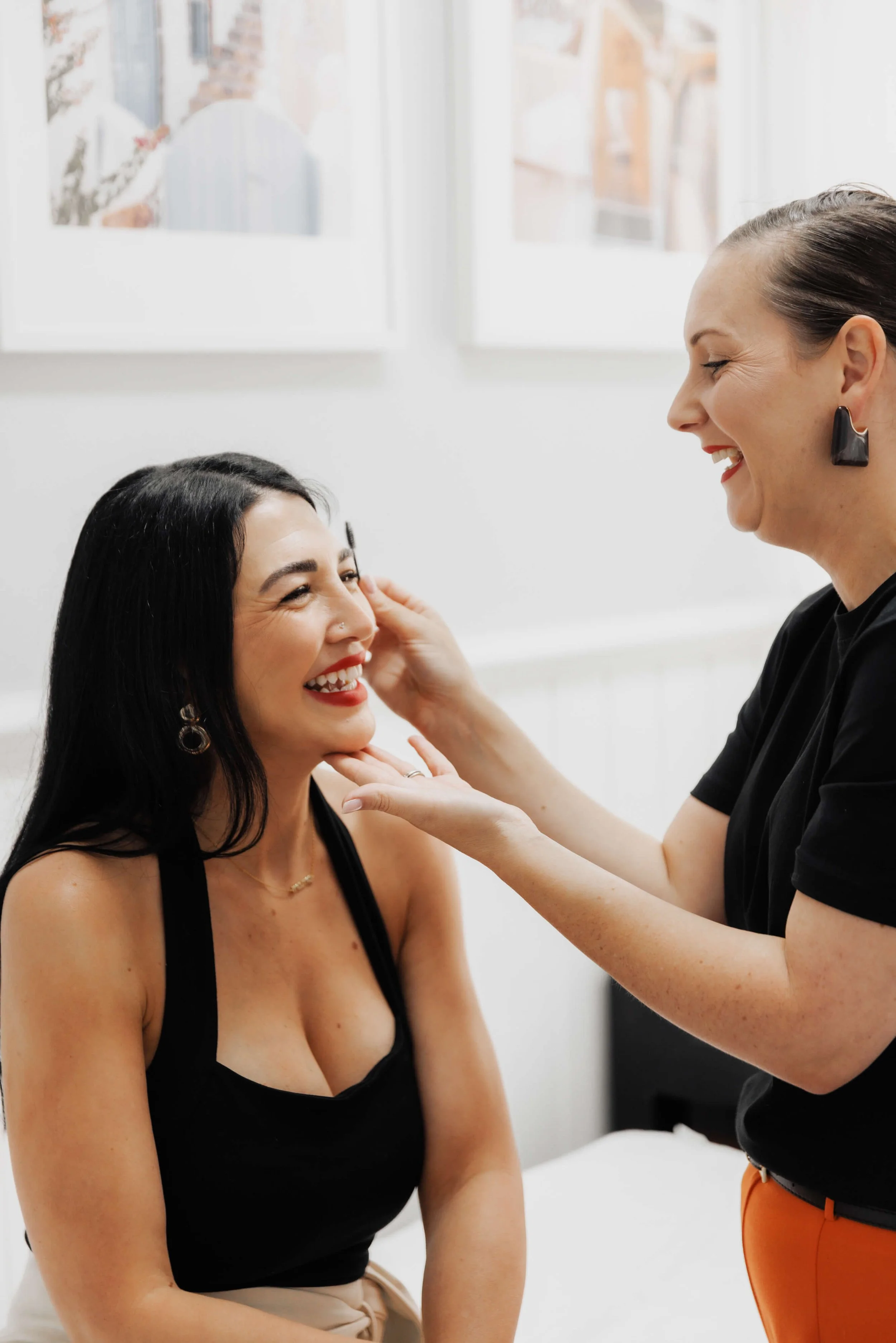 A woman with dark hair and a nose piercing gets her eyebrows waxed by a professional with short brown hair, wearing a black top and orange pants, in a bright room.