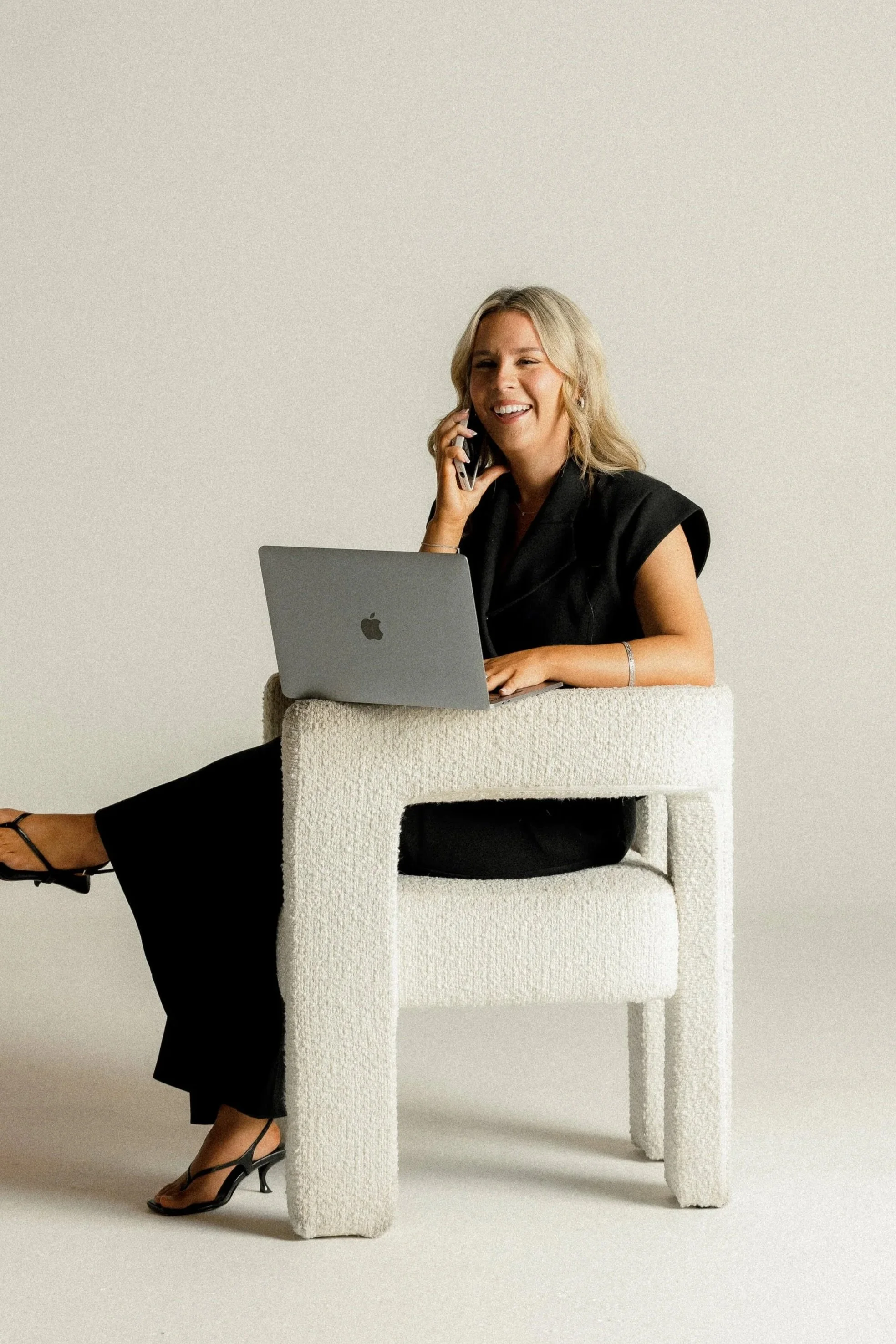 Ciara sitting on a white textured chair with a laptop on her lap, talking on a phone.