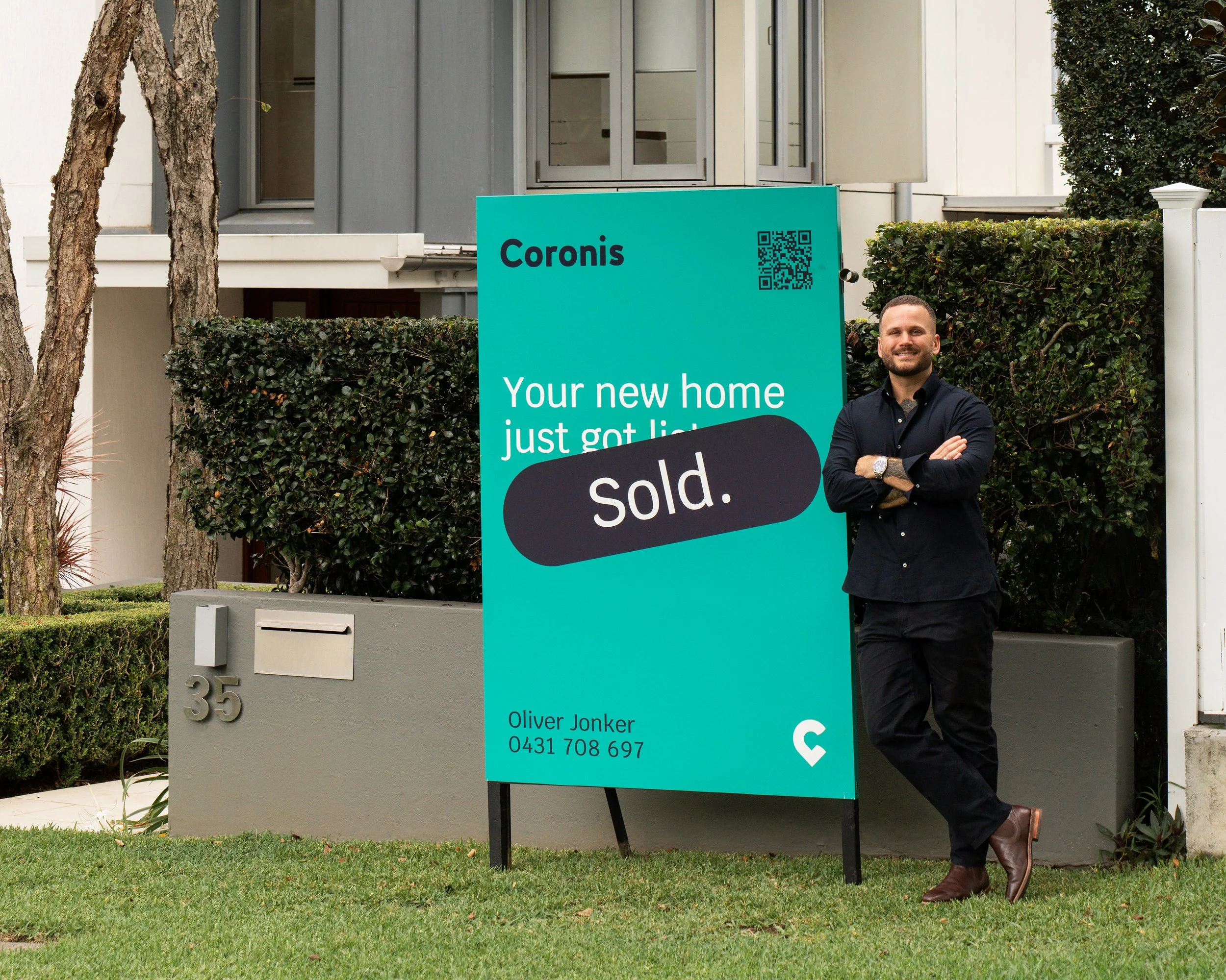 A man in black clothing with crossed arms smiling next to a teal real estate sign that reads 'Coronis' and 'sold'. The sign is in a residential front yard with trees, bushes, and a modern house in the background.