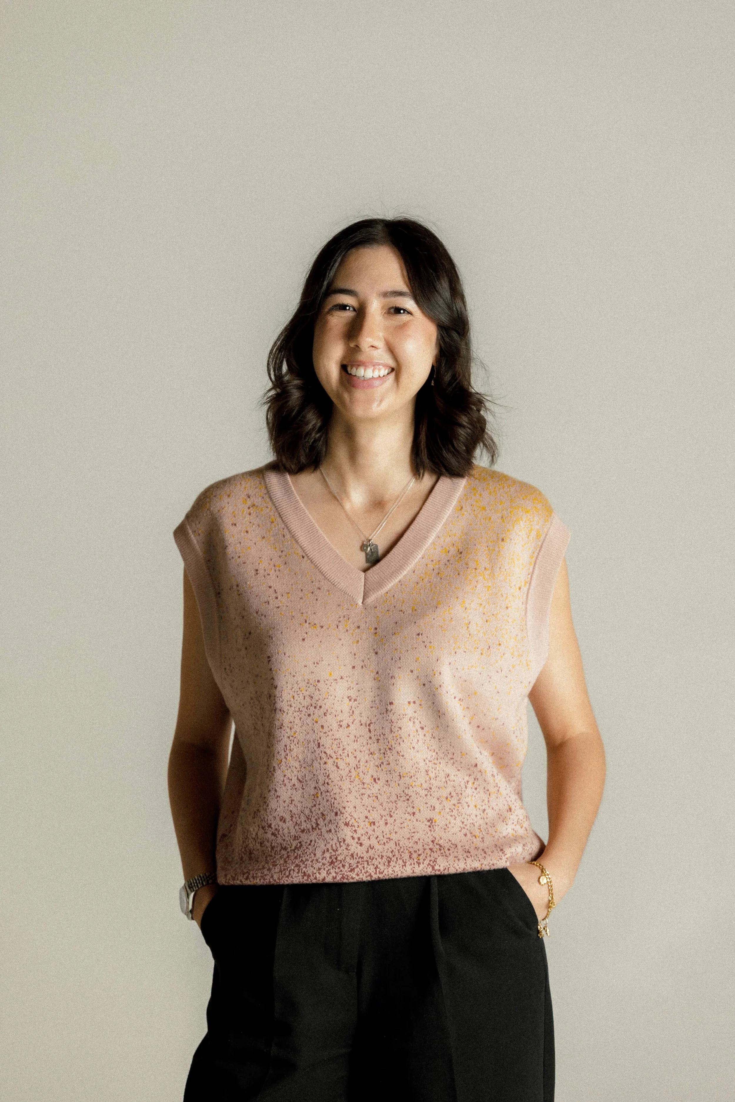 A woman with shoulder-length dark hair, smiling, wearing a sleeveless pink and yellow speckled sweater, black pants, a necklace, watch, and bracelet, standing against a plain light-colored background.