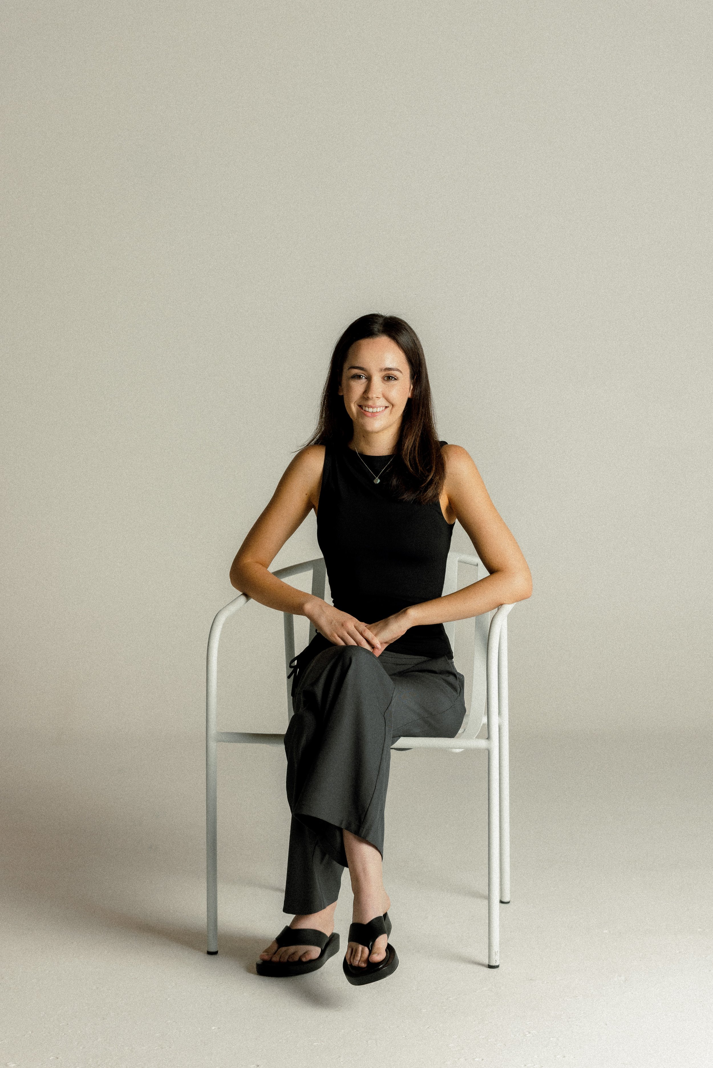A young woman sitting on a white chair against a plain beige background, wearing a black sleeveless top, gray pants, black sandals, and a necklace, smiling at the camera.