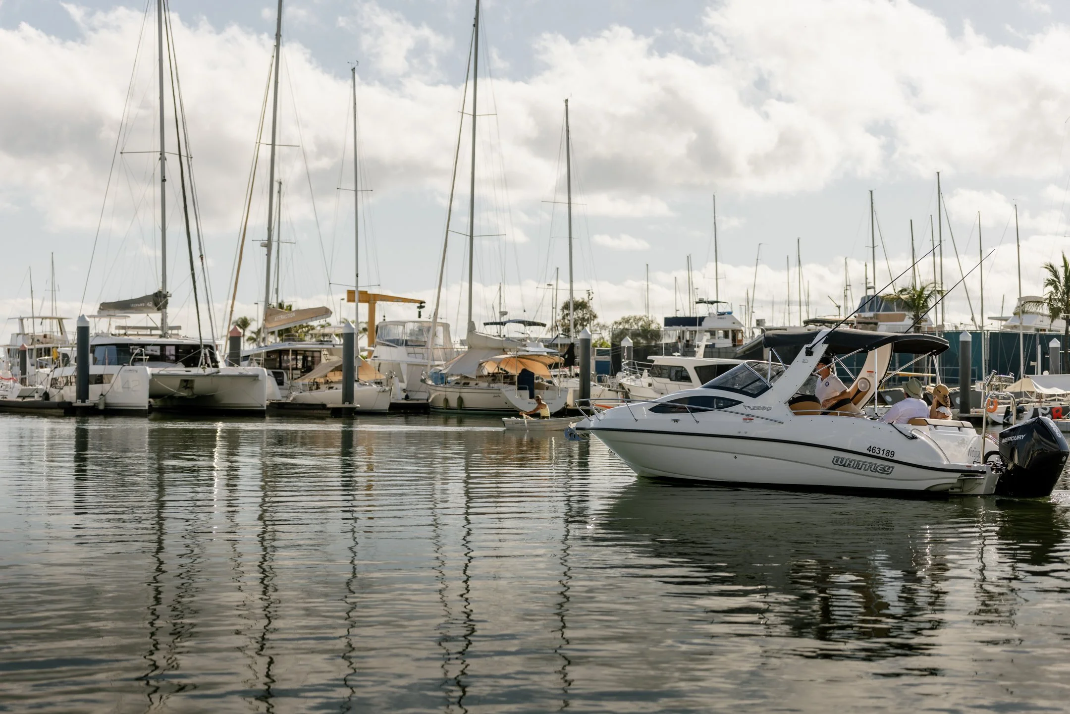 A marina with numerous sailboats and motorboats docked, with people on some boats, and a cloudy sky reflected on the water.