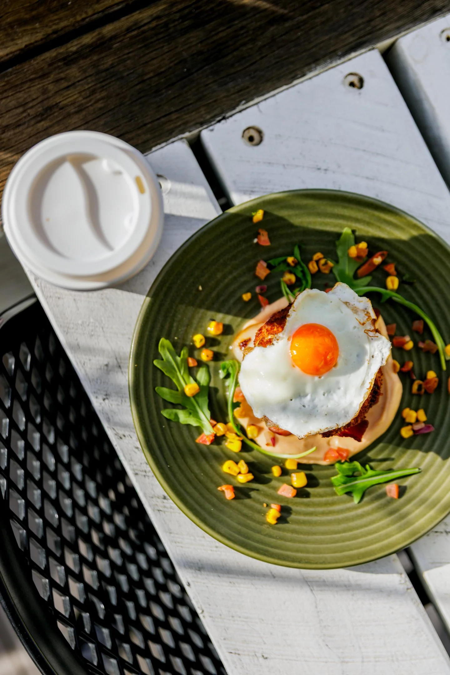 A plate with a sunny side up egg on top of a piece of toast, garnished with fresh greens and small colorful vegetable pieces, on a white wooden table with a cup of coffee nearby.