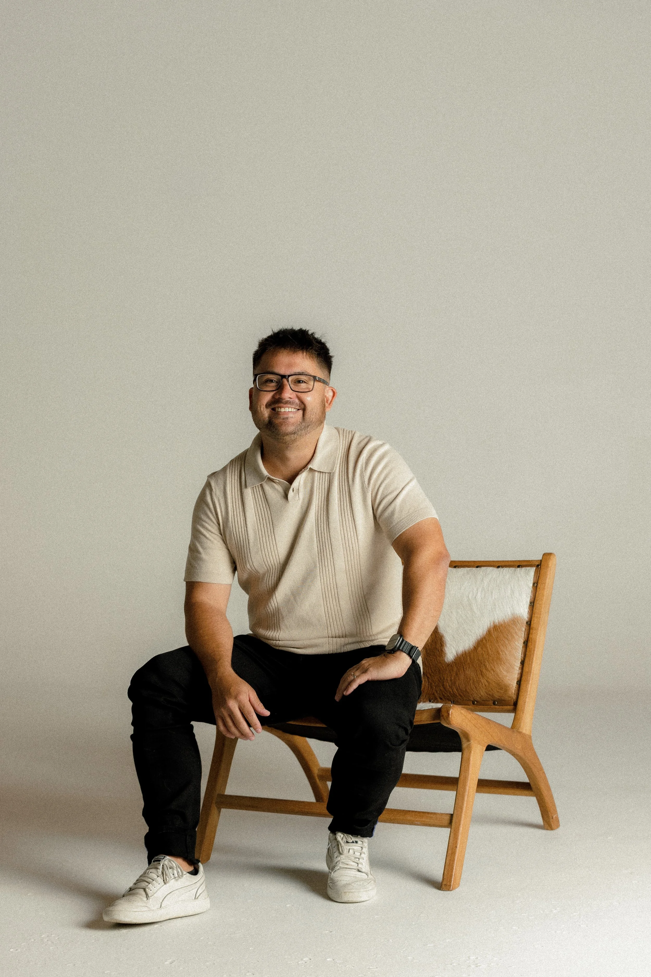 A smiling man with glasses, wearing a beige short-sleeve polo shirt and black pants, sitting on a wooden chair with a fur accent, against a neutral background.