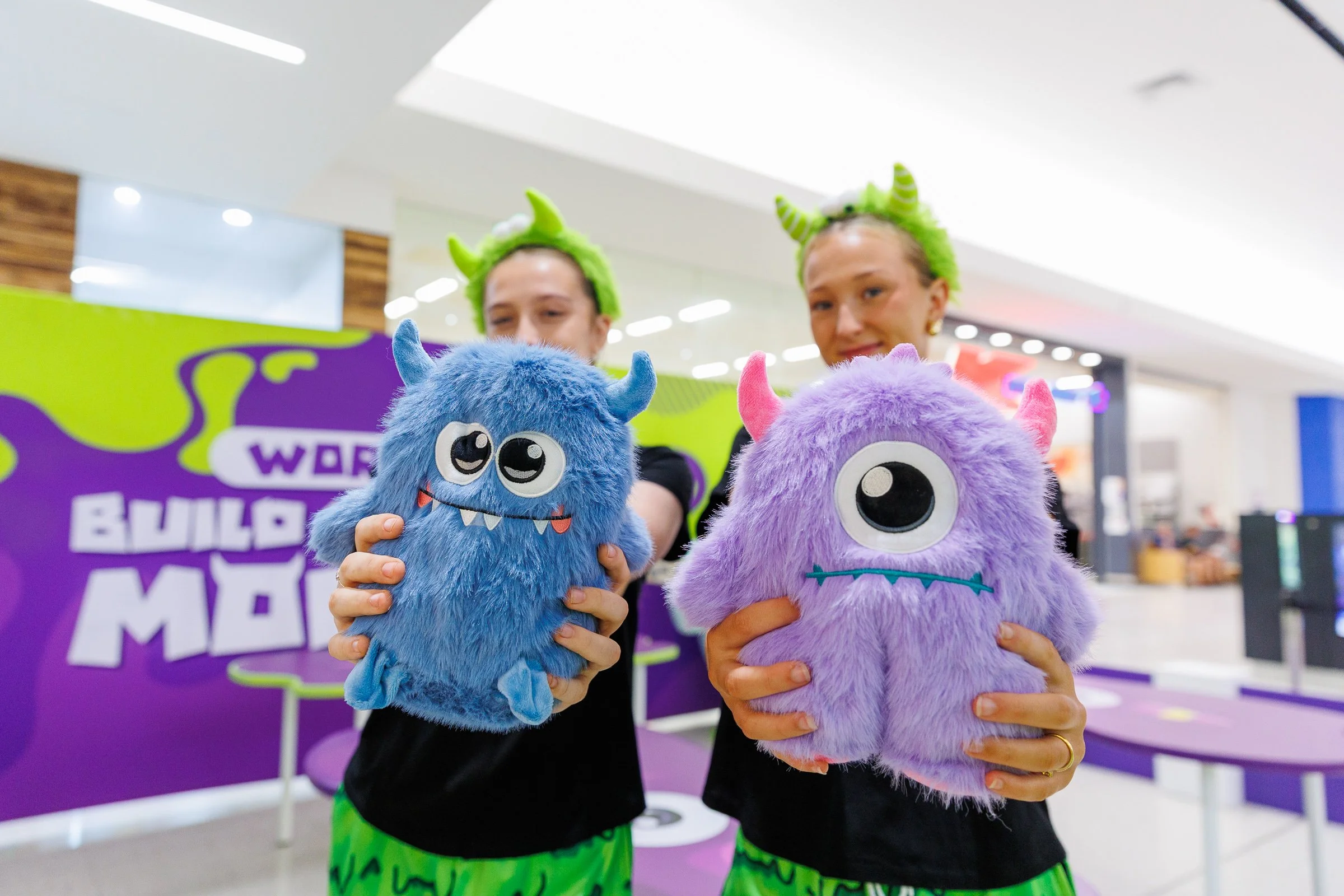Two people holding colorful furry monster plush toys, one blue and one purple, inside a shopping mall with a vibrant purple and green backdrop.