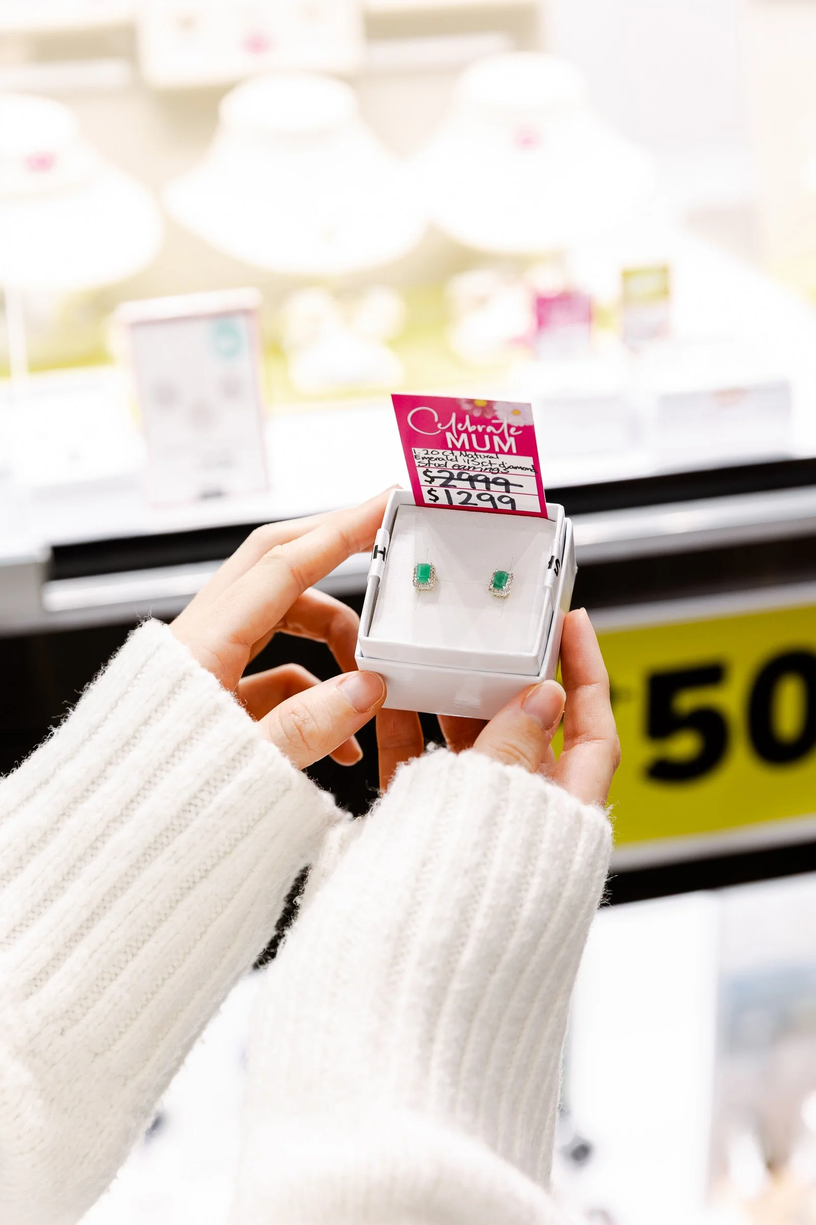 Close-up of a pair of green gemstone earrings in a jewellry box, with a pink sale sign held by a person wearing a white knitted sweater in a retail store.