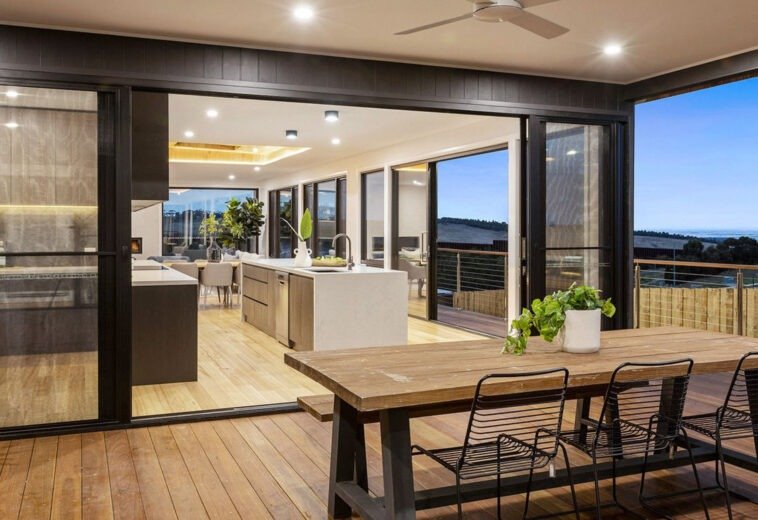 A modern house's outdoor entertaining space with wooden floors and a large sliding glass door that opens onto a sophisticated kitchen and dining room.