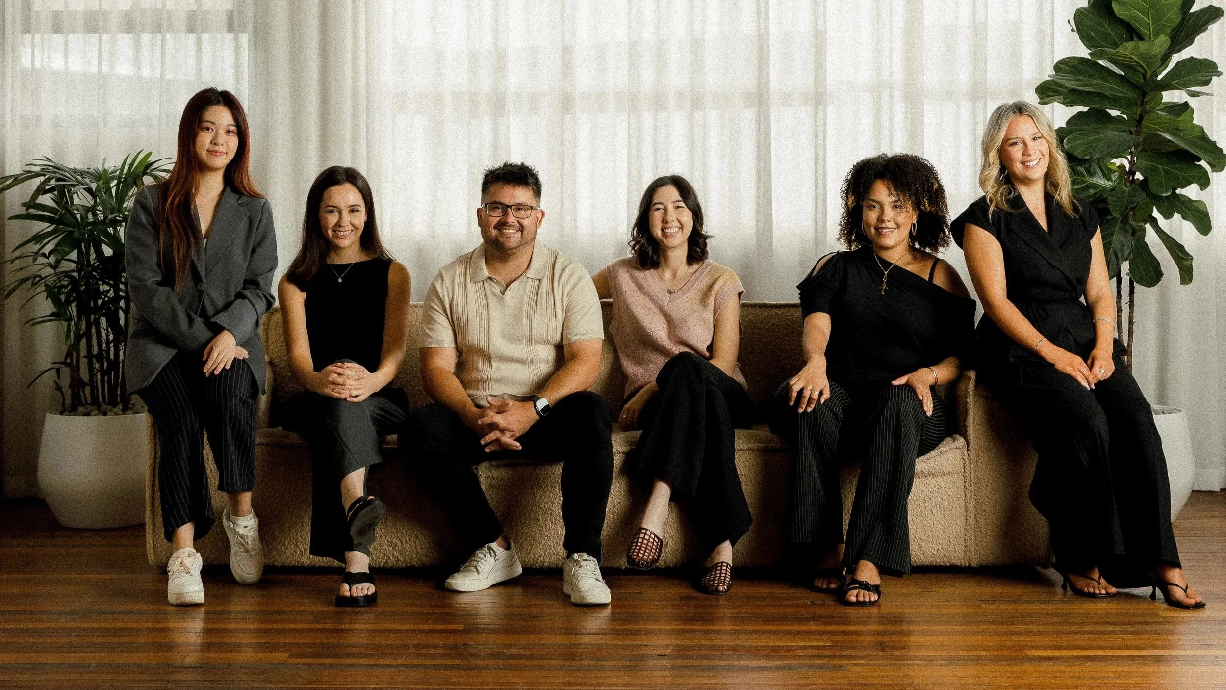 The Allies team smiling sitting on a beige couch with a curtain and plants behind.