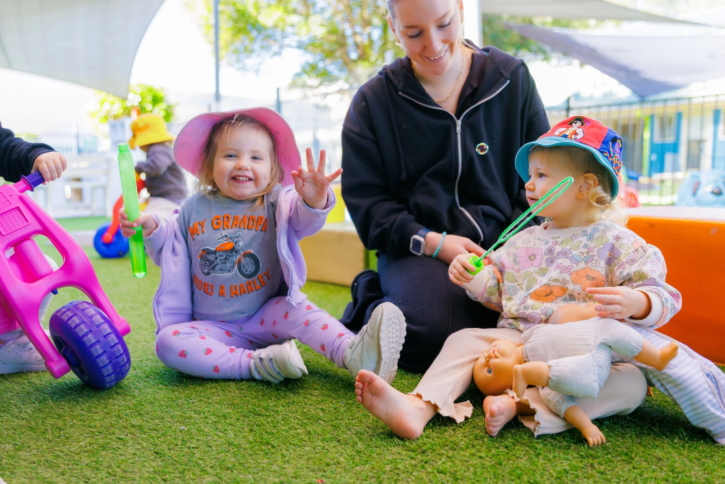 A group of young children and a woman playing outside on artificial grass. One girl is holding a bubble stick, another girl is sitting next to a baby doll, and a woman is smiling and sitting with them. Toys and trees are visible in the background.