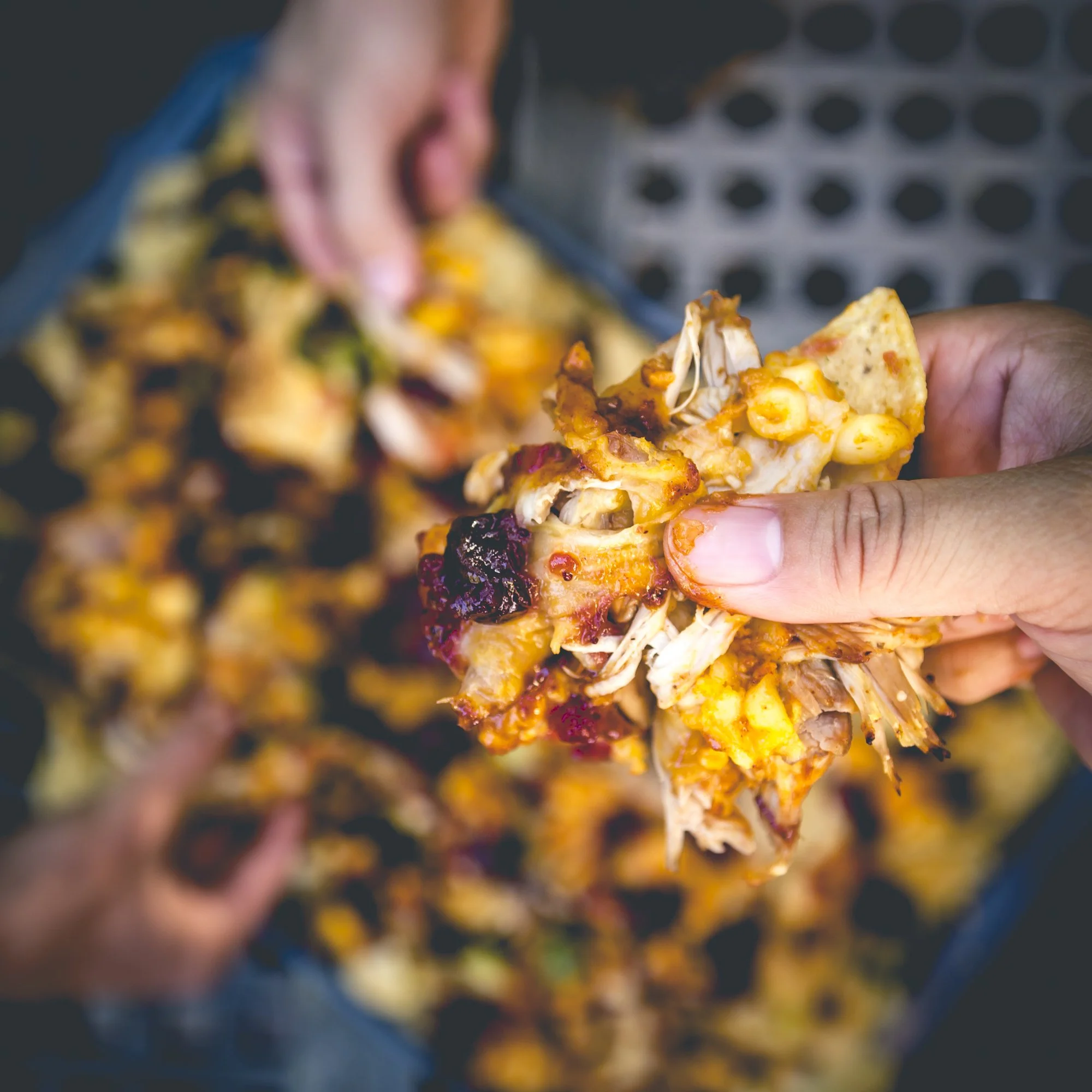 Person holding a piece of pizza with cheese, chicken, and toppings, with a tray of more pizza slices in the background.