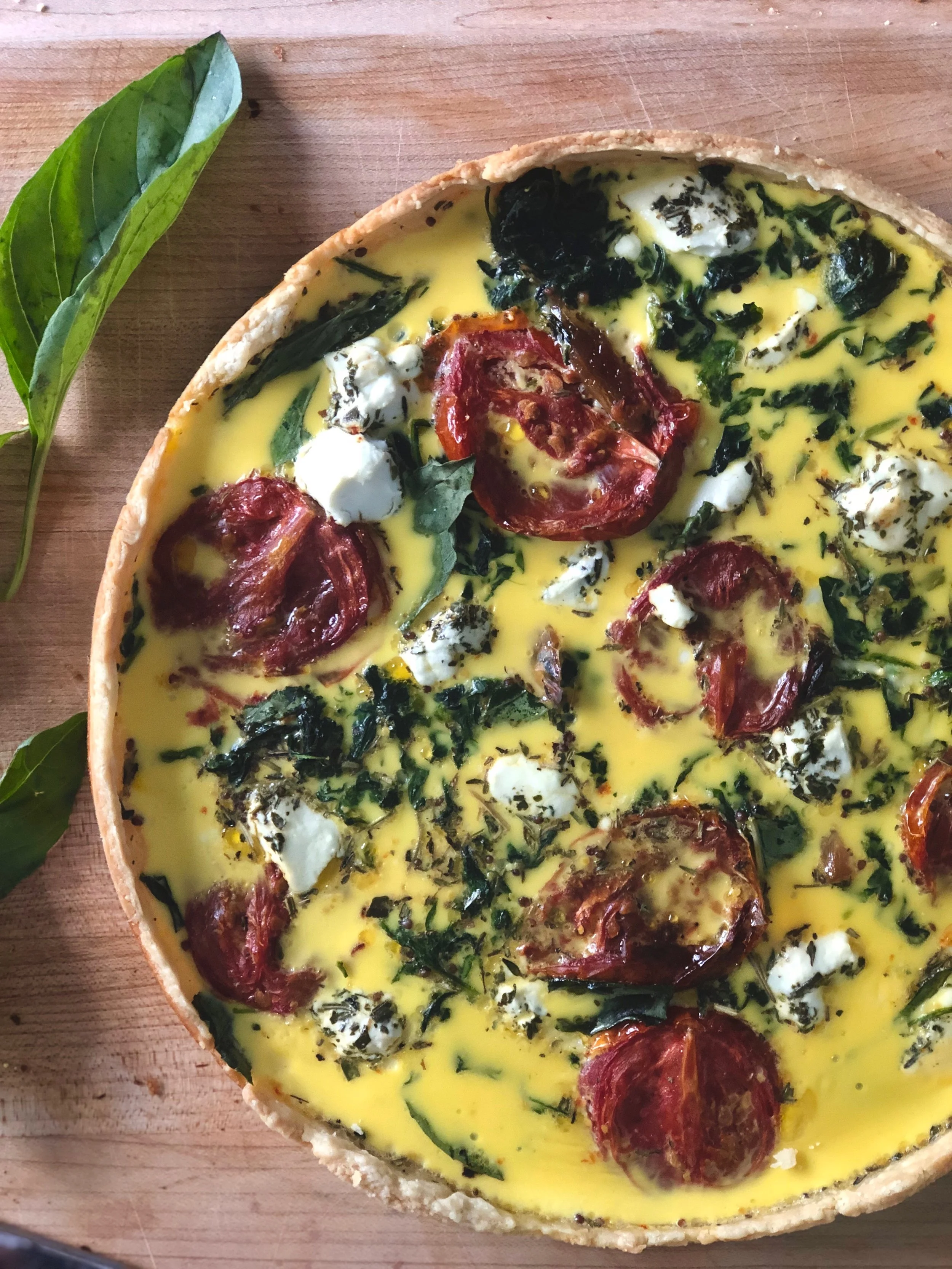Quiche with cherry tomatoes, spinach, and dollops of cream, on a wooden surface with green leaves nearby.