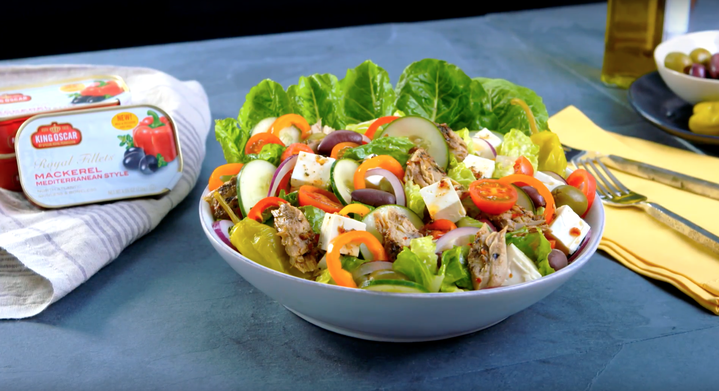 Bowl of fresh Mediterranean-style salad with lettuce, tomatoes, cucumbers, olives, red onion, feta cheese, and chicken on a dark table next to a yellow napkin and silverware.
