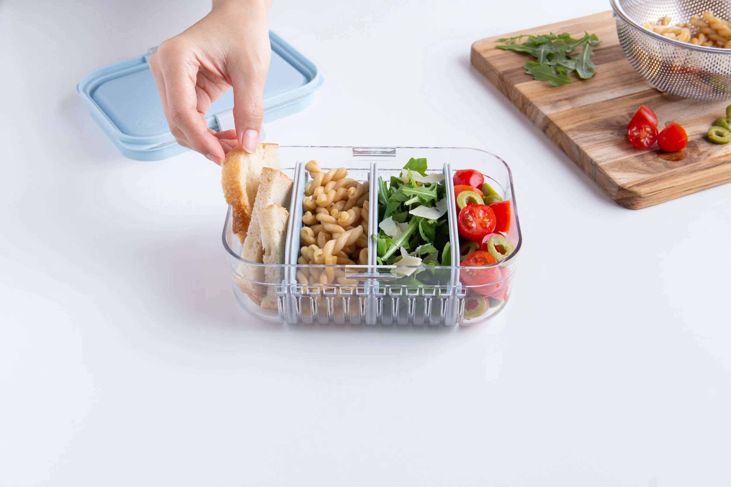 A hand packing bread, pasta, and salad ingredients into a divided food storage container on a white surface.