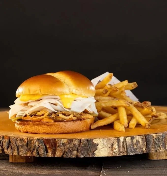 A chicken sandwich with cheese, coleslaw, and onion rings on a wooden serving board, along with French fries.