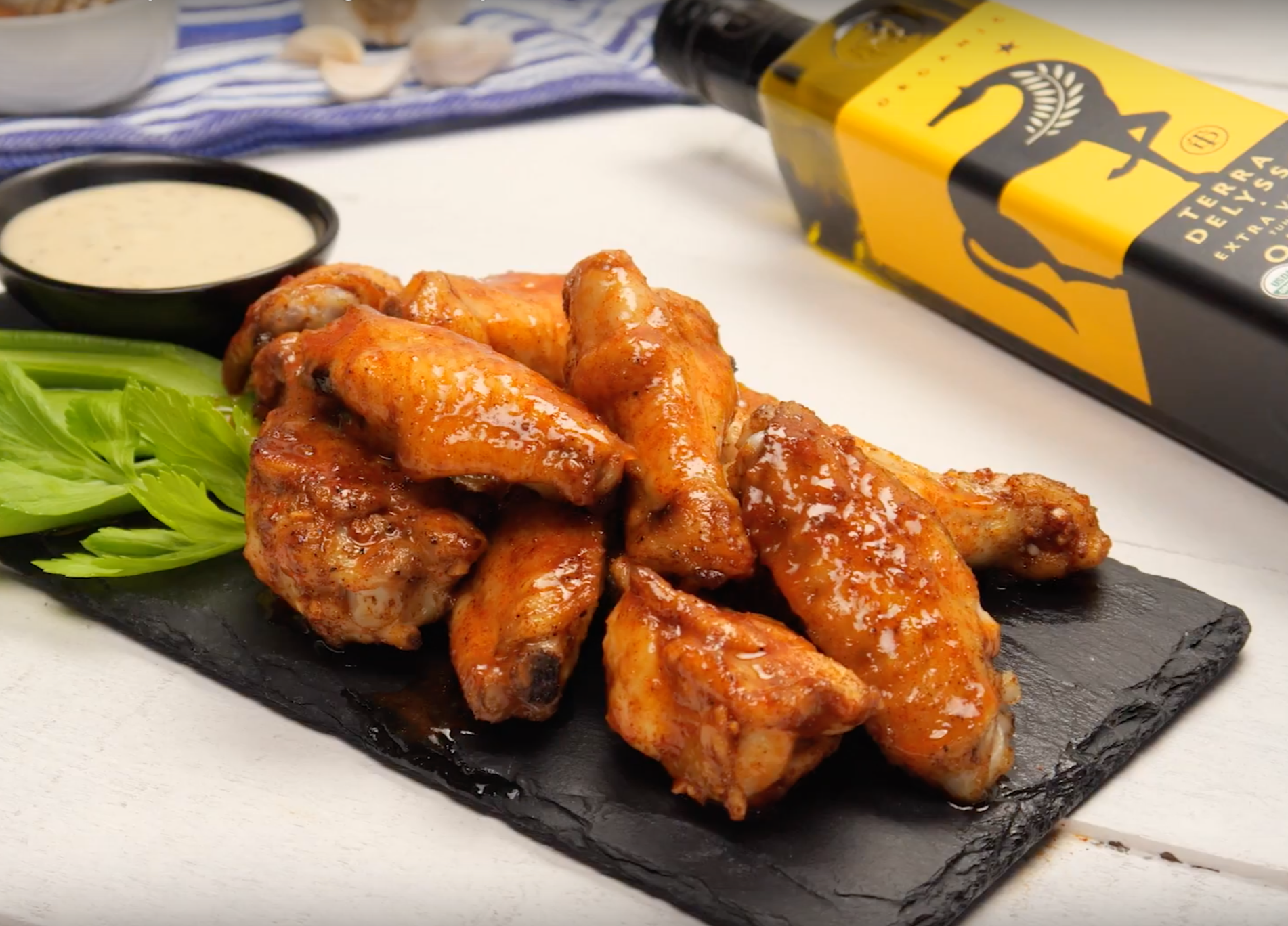 A slate platter of glazed chicken wings with celery stalks, a small bowl of dipping sauce, and a bottle of Terraz Delyssa extra virgin olive oil in the background.