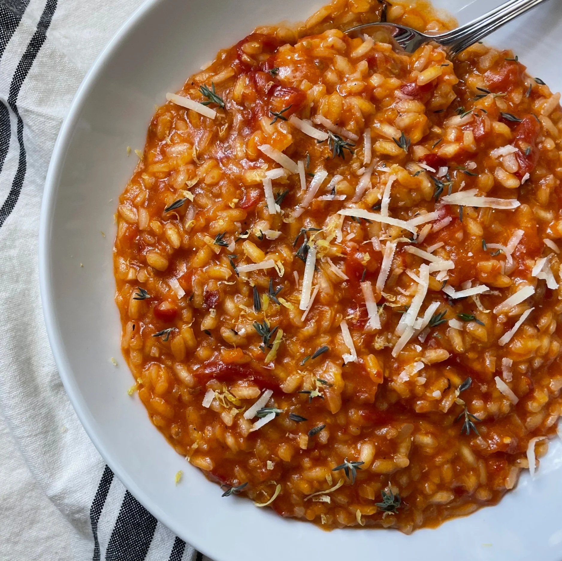 A white bowl filled with tomato-based risotto garnished with shredded cheese and fresh herbs, served on a striped cloth napkin.