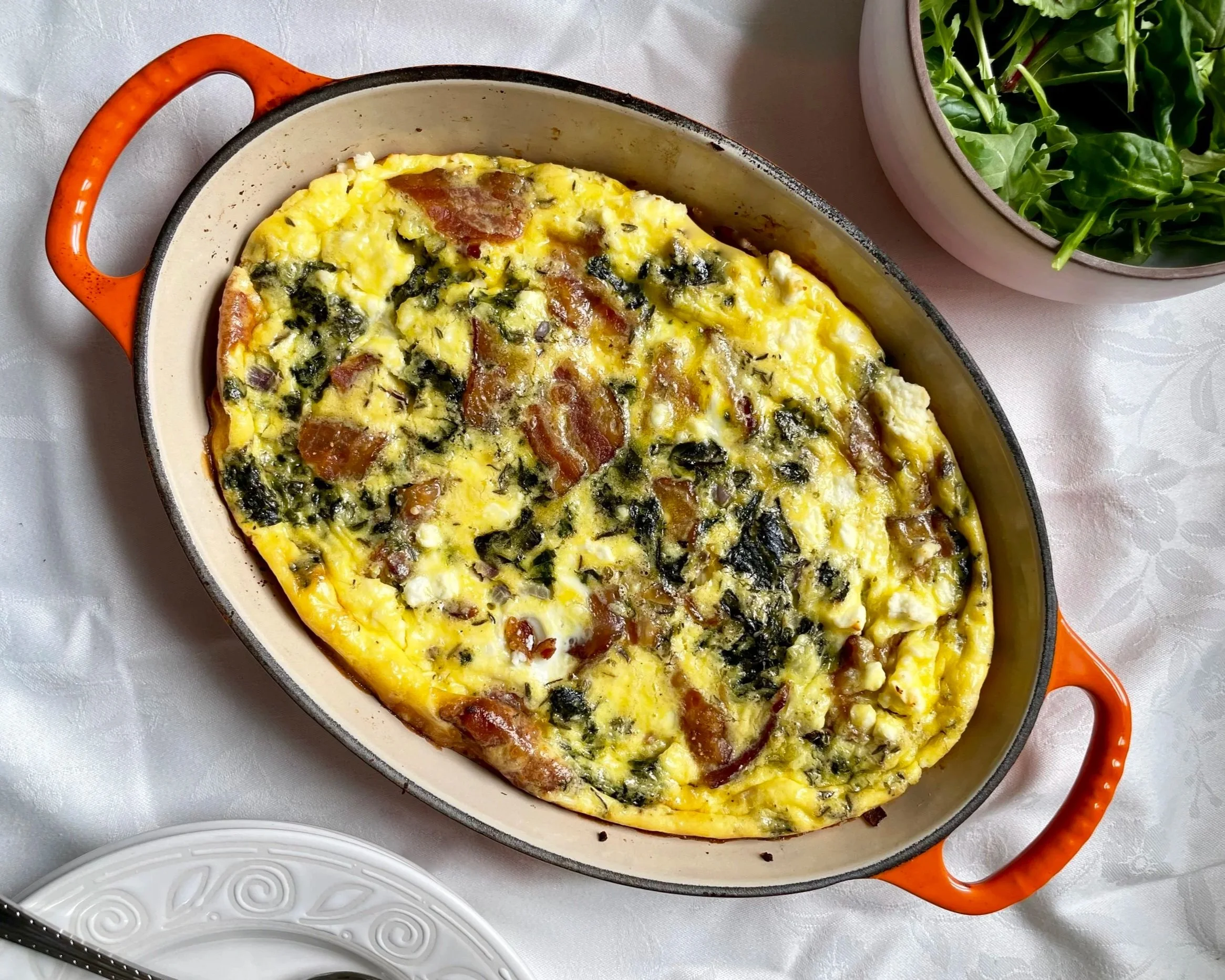 Baked frittata with eggs, spinach, and bacon in a ceramic baking dish on a table with a bowl of salad.