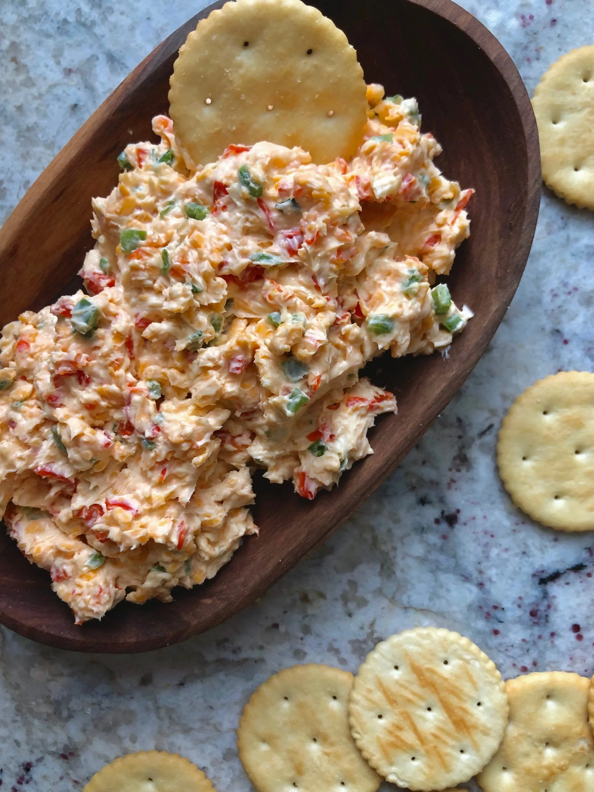 A wooden bowl contains chicken salad with chopped vegetables, nearby are round crackers on a marble surface.