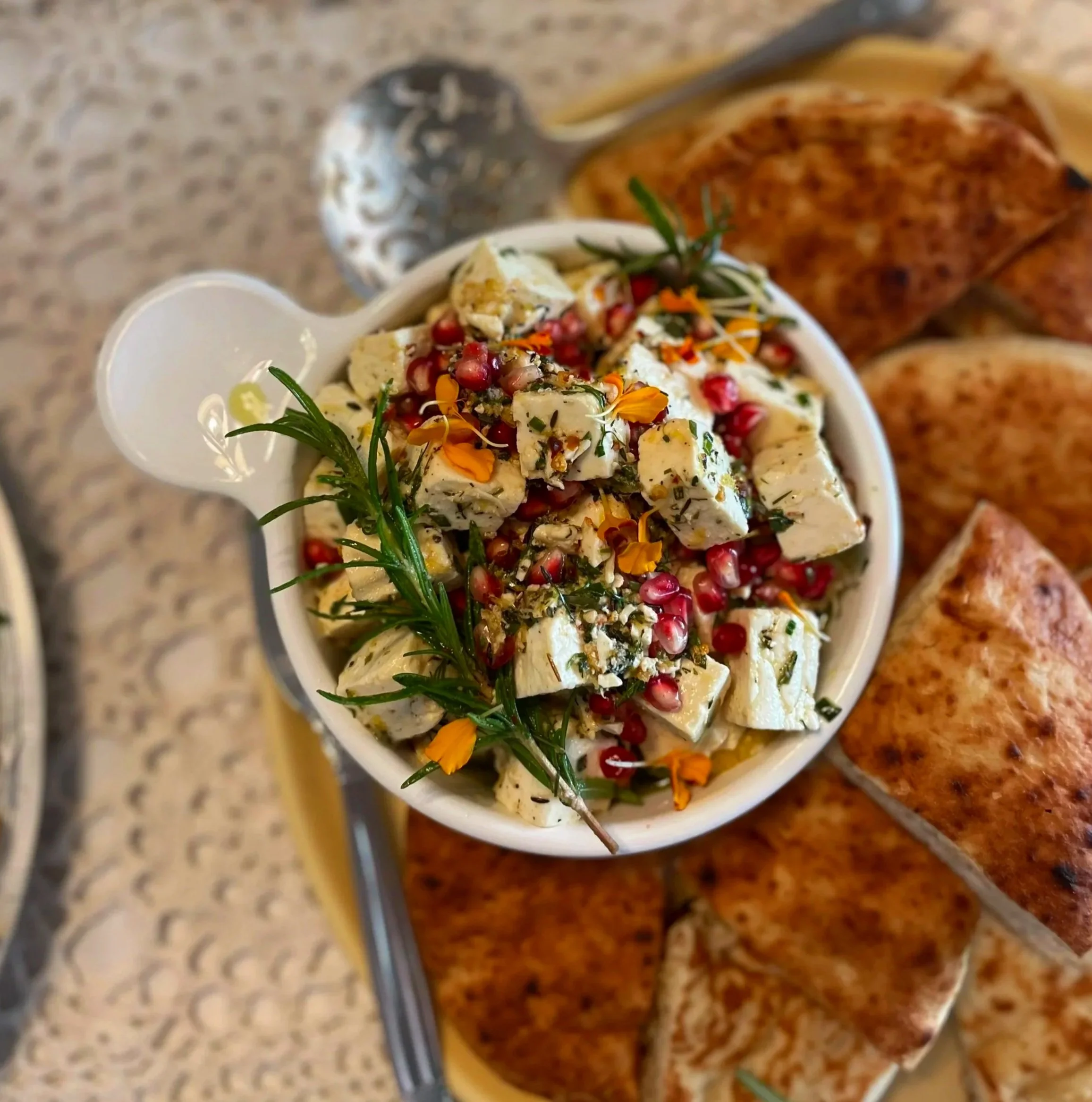 A bowl of tofu salad garnished with herbs and pomegranate seeds, with toasted bread slices on a tray in the background.