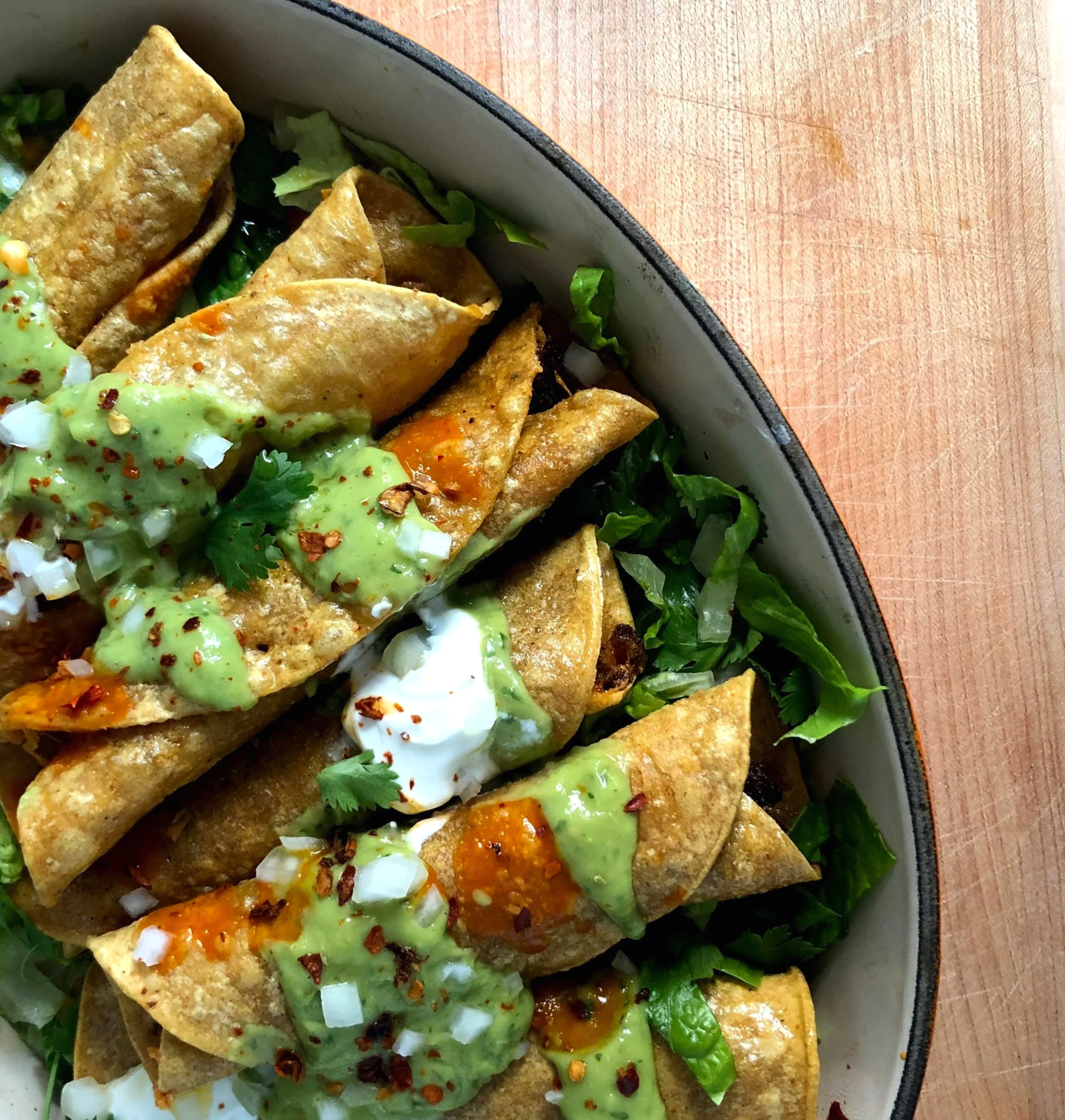 Close-up of a plate of taquitos topped with green sauce, sour cream, chopped onions, cilantro, and red chili flakes, with chopped lettuce underneath.