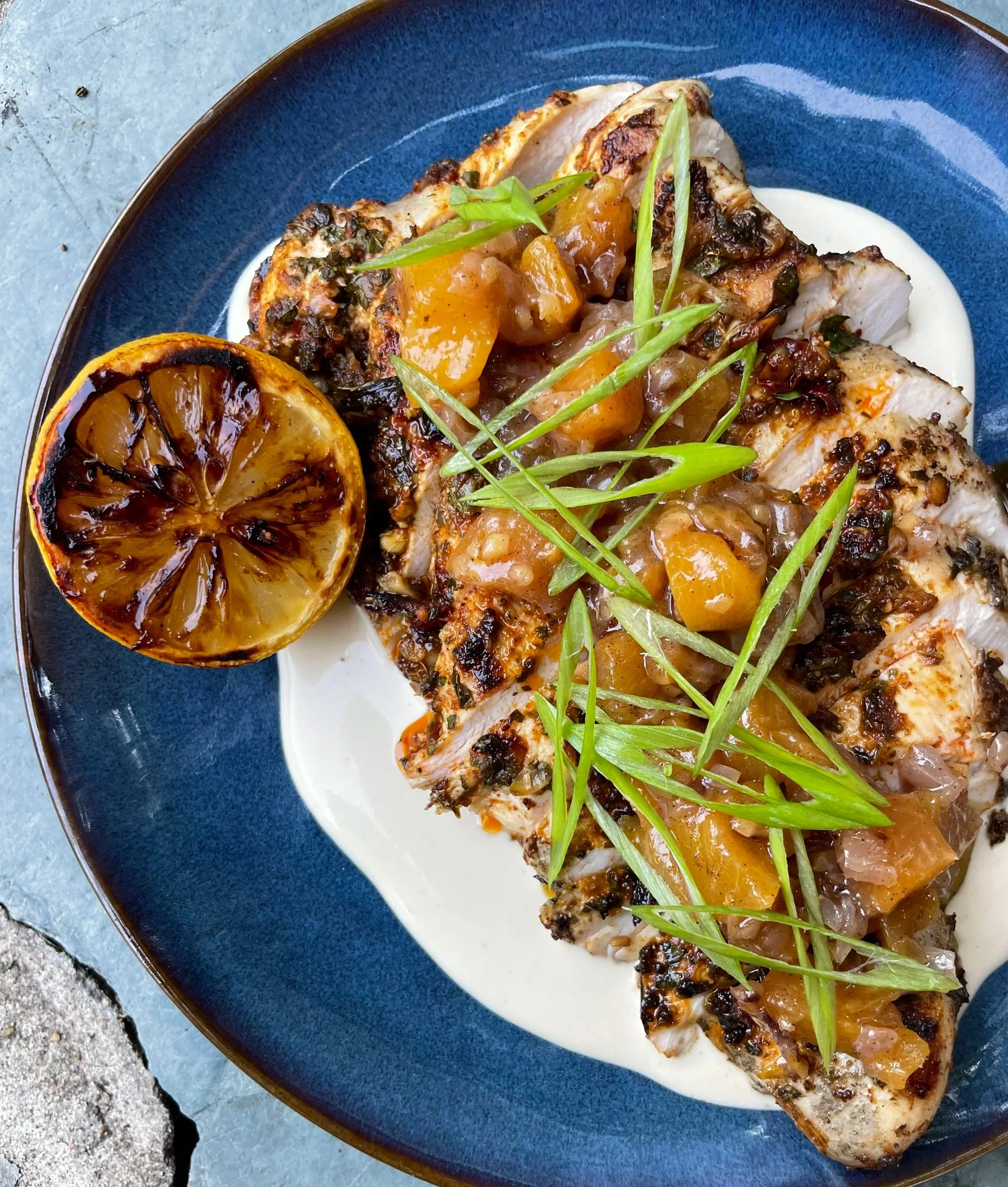 Grilled chicken breast topped with a fruit chutney garnished with green onion slices, served on a blue plate with a roasted lemon half on the side.