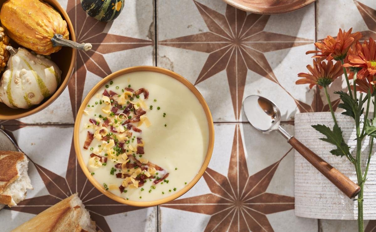 A bowl of creamy soup garnished with chopped chives, diced ham, and shredded cheese, placed on a patterned tile table. Surrounding the bowl are small decorative gourds, a bunch of orange flowers, a spoon, and some bread.