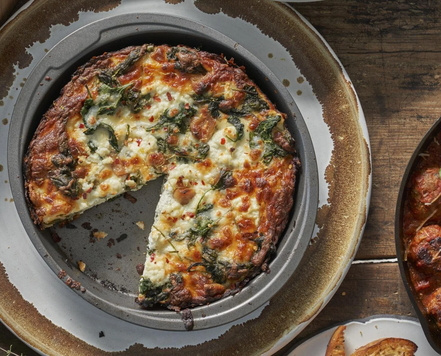 A veggie pizza with cheese, spinach, and red pepper flakes on a dark round pizza pan. Part of another dish is visible on the right edge on a wooden table.