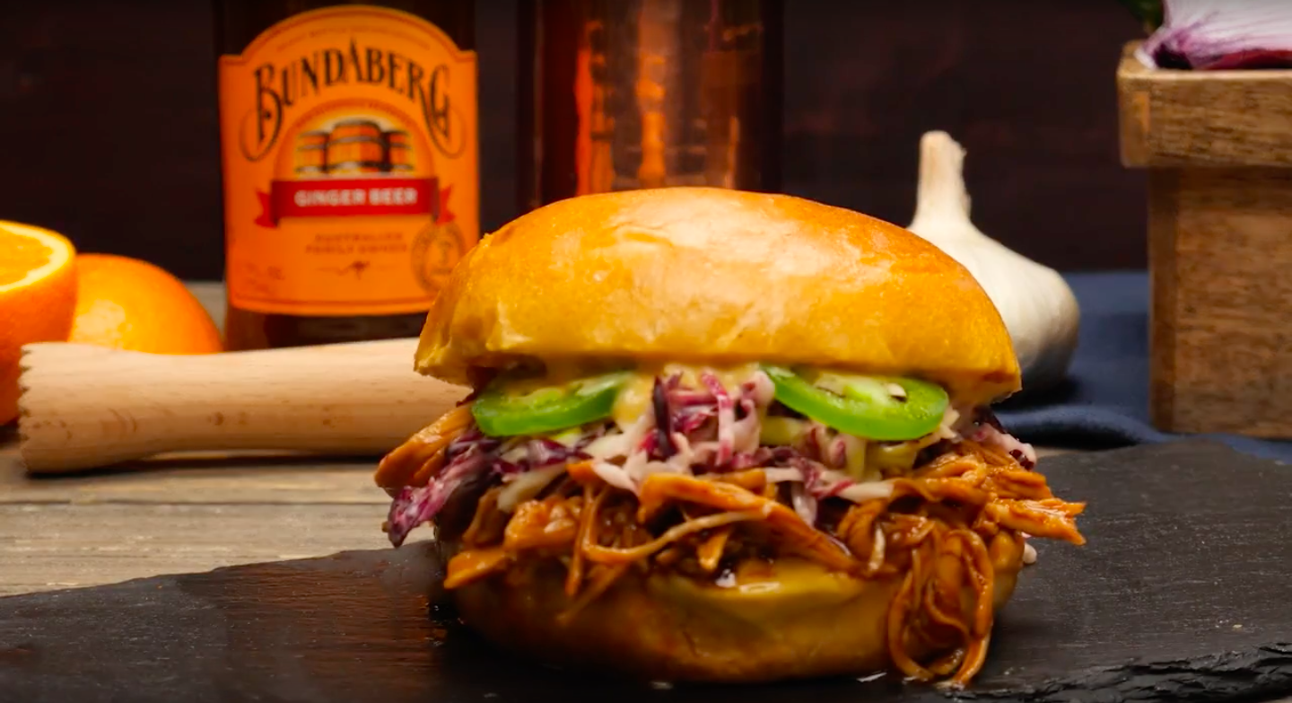 Pulled pork sandwich with coleslaw and pickles on a bun, placed on a black slate serving board. In the background, there are oranges, a jar of ginger beer labeled 'Bundaberg', a garlic bulb, and a wooden box.