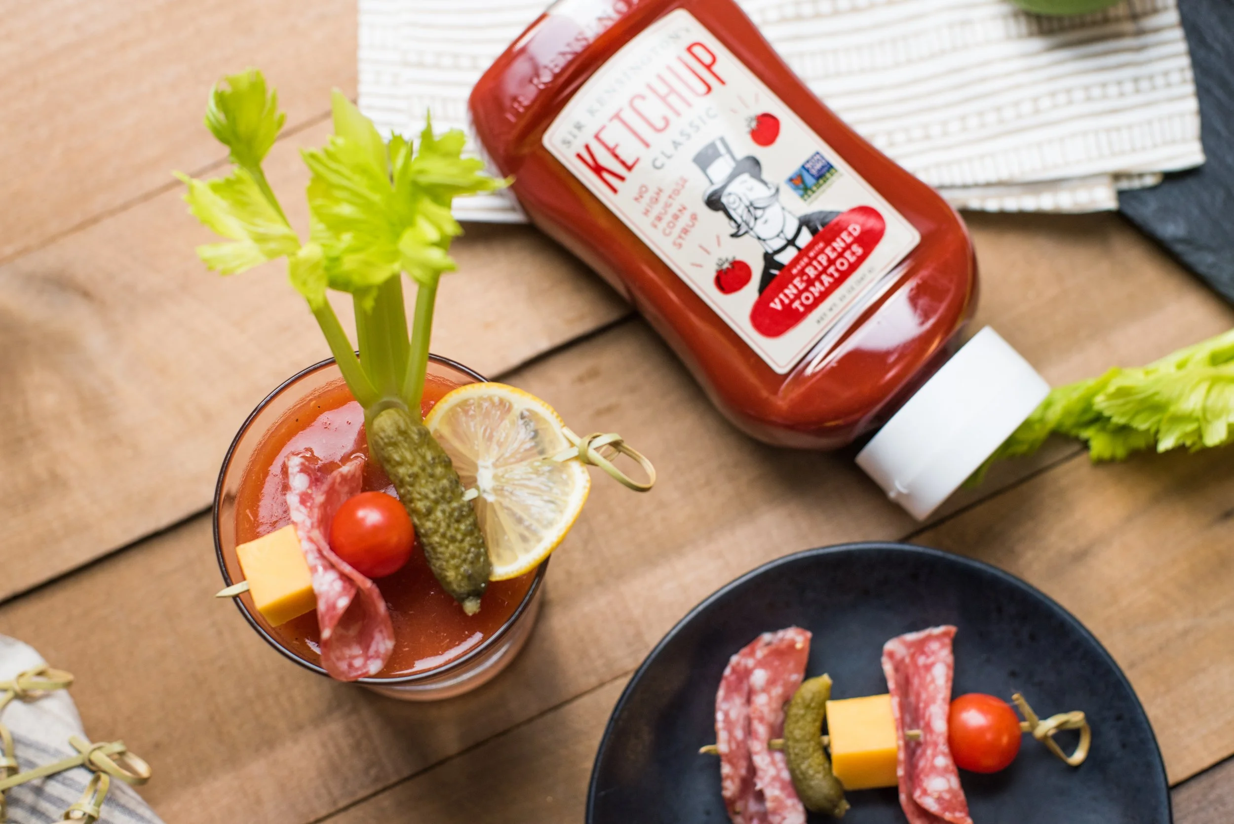 A glass filled with red cocktail garnished with a lemon slice, cherry tomato, cheese cube, salami, pickle, and celery stalk, placed on a wooden table next to a black plate with salami, cheese, and cherry tomato, and a bottle of ketchup labeled ketchu