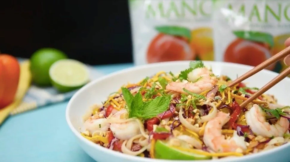 A bowl of shrimp and noodle salad with lime and herbs, chopsticks, and fresh fruit in the background.