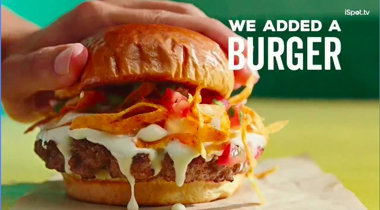 A close-up of a burger with a bun, meat patty, melted cheese, shredded lettuce, tomato slices, and other toppings, with hands holding the bun. Text on the image reads, 'We added a burger.'