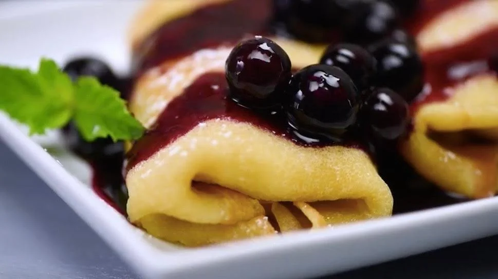 Close-up of crepes topped with blueberry compote and powdered sugar on a white plate.