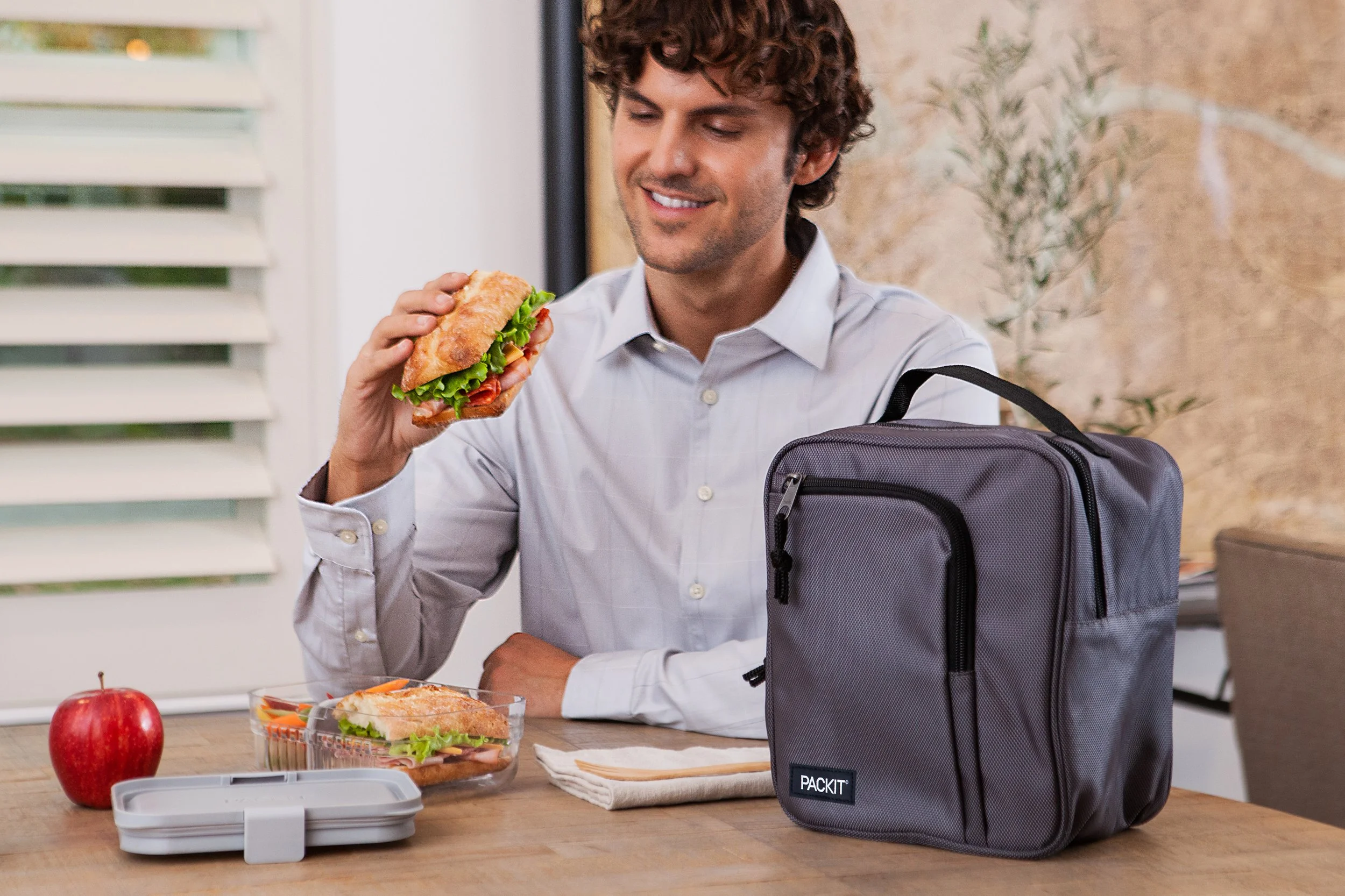 A man sitting at a wooden table with a gray lunch bag, an apple, and a sandwich container with a sandwich inside. He is holding a sandwich in his right hand and smiling. The background shows a window with blinds and a plant.