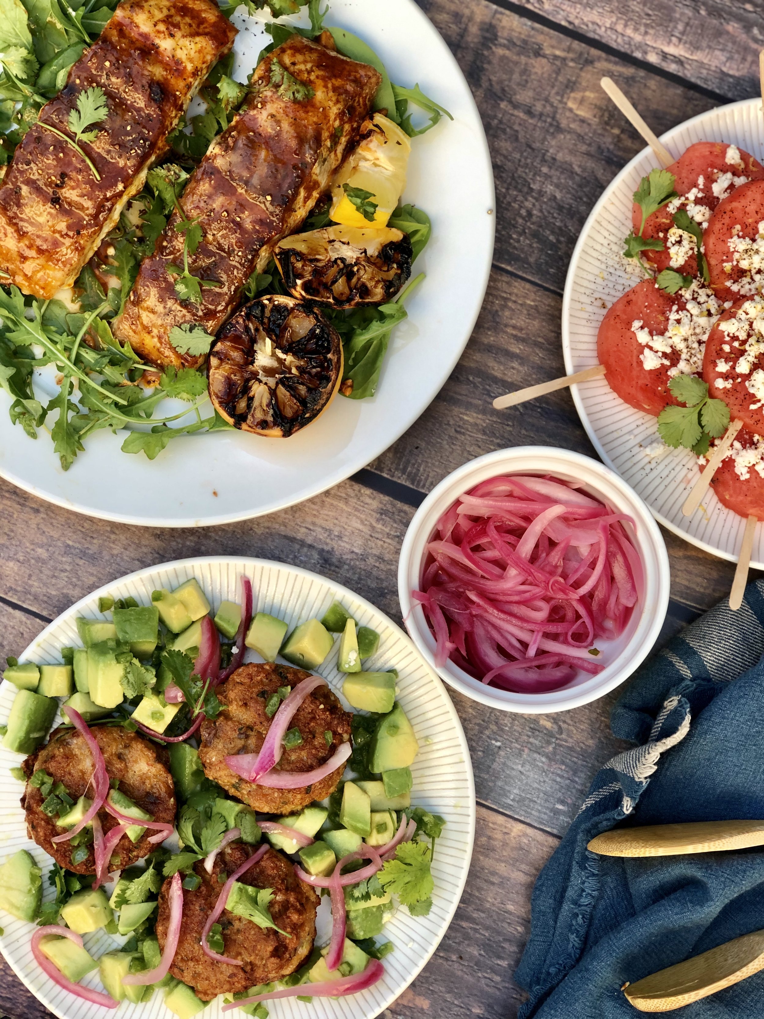 Variety of colorful Mexican dishes on plates, including grilled fish with charred lemon and greens, avocado and fried meatballs with onions, tomato skewers with cheese and herbs, and pickled red onions, all on a rustic wooden table.