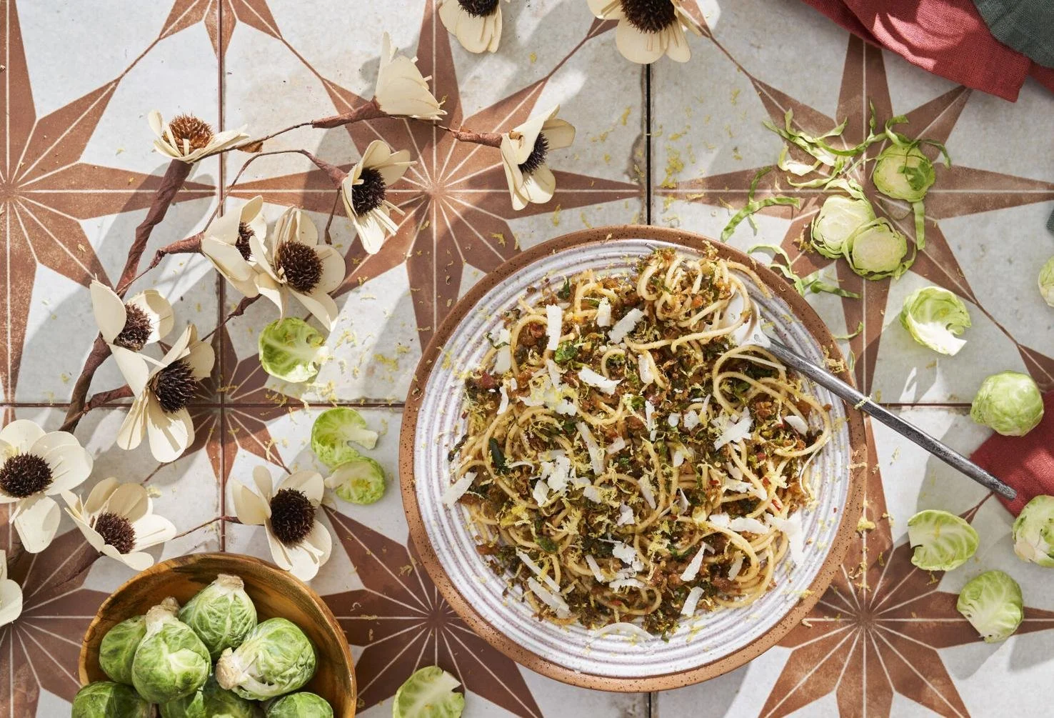 A bowl of spaghetti pasta with sauce, cheese, and herbs on a decorative tile surface surrounded by Brussels sprouts and a floral garland.