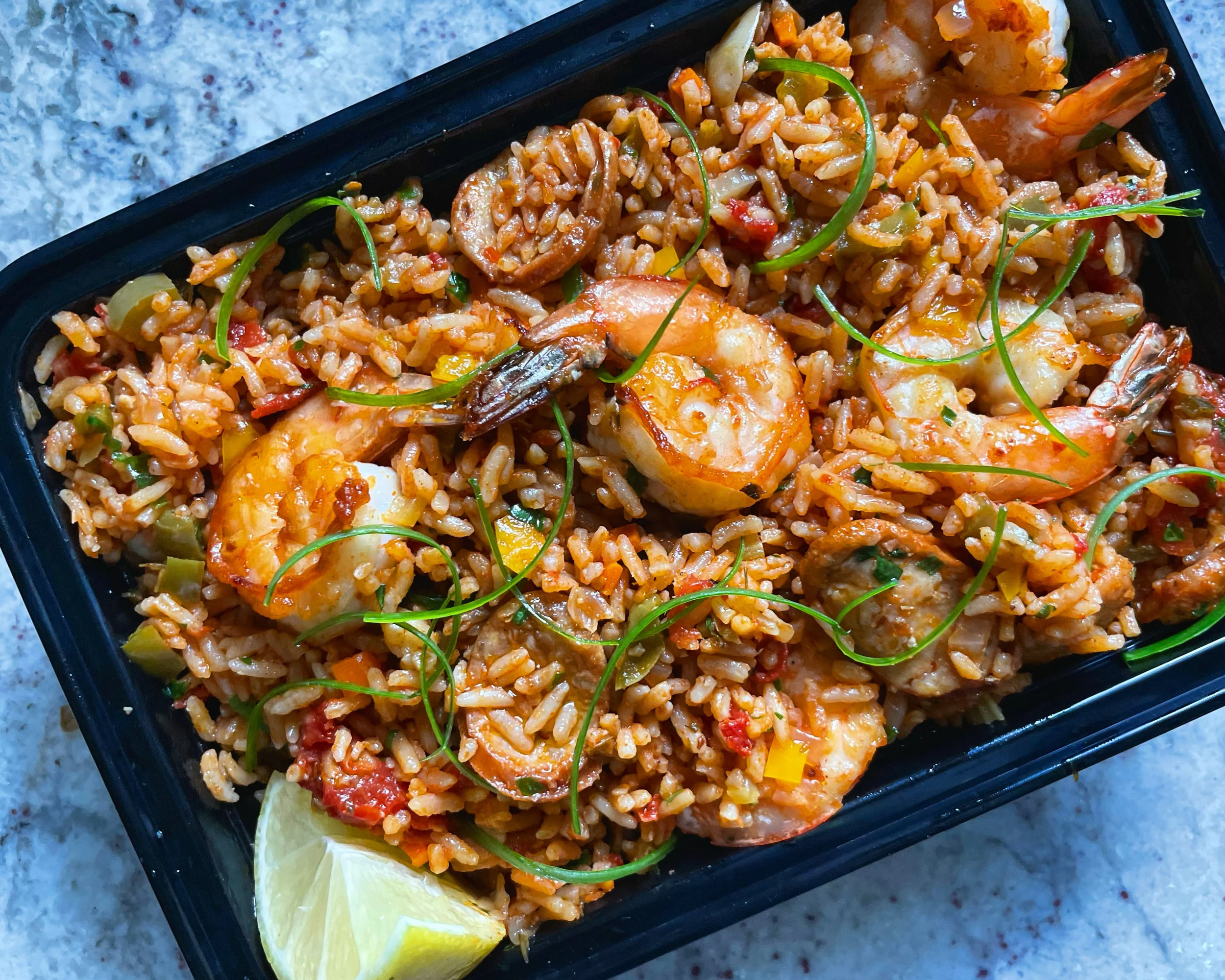 A black container of cooked shrimp and rice with vegetables, garnished with green herbs and lemon wedges.
