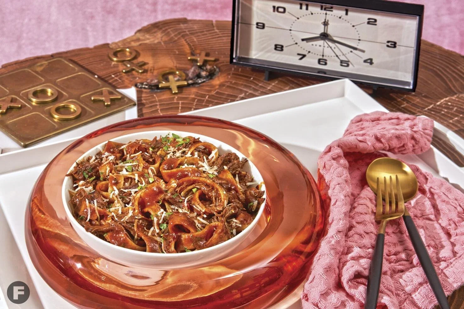 A bowl of pasta with tomato sauce and grated cheese on a red glass plate, set on a white tray with a pink cloth napkin, gold fork and spoon. In the background, a checkered board, a clock, and some jewelry on a wooden surface.