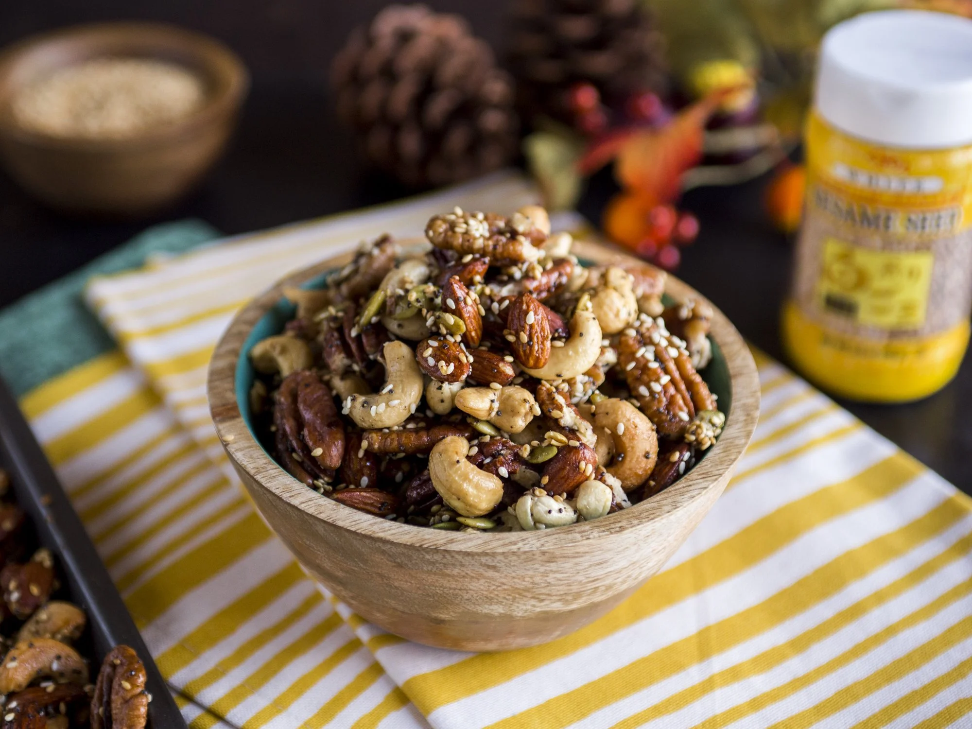 A wooden bowl filled with mixed nuts including cashews, almonds, pecans, and pumpkin seeds, topped with sesame seeds, placed on a yellow and white striped cloth. In the background, there are some pinecones, a jar of mustard seed, and a decorative aut