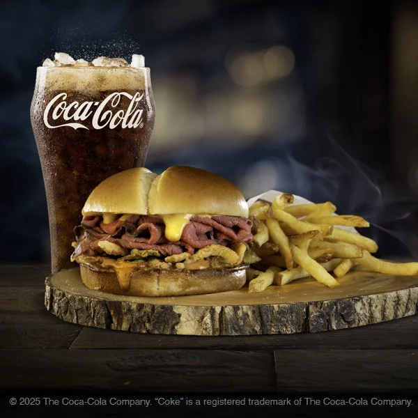 A cheeseburger with beef patty, cheese, bacon, and steak inside a bun, served with French fries, a tall Coca-Cola, and a wooden serving board.