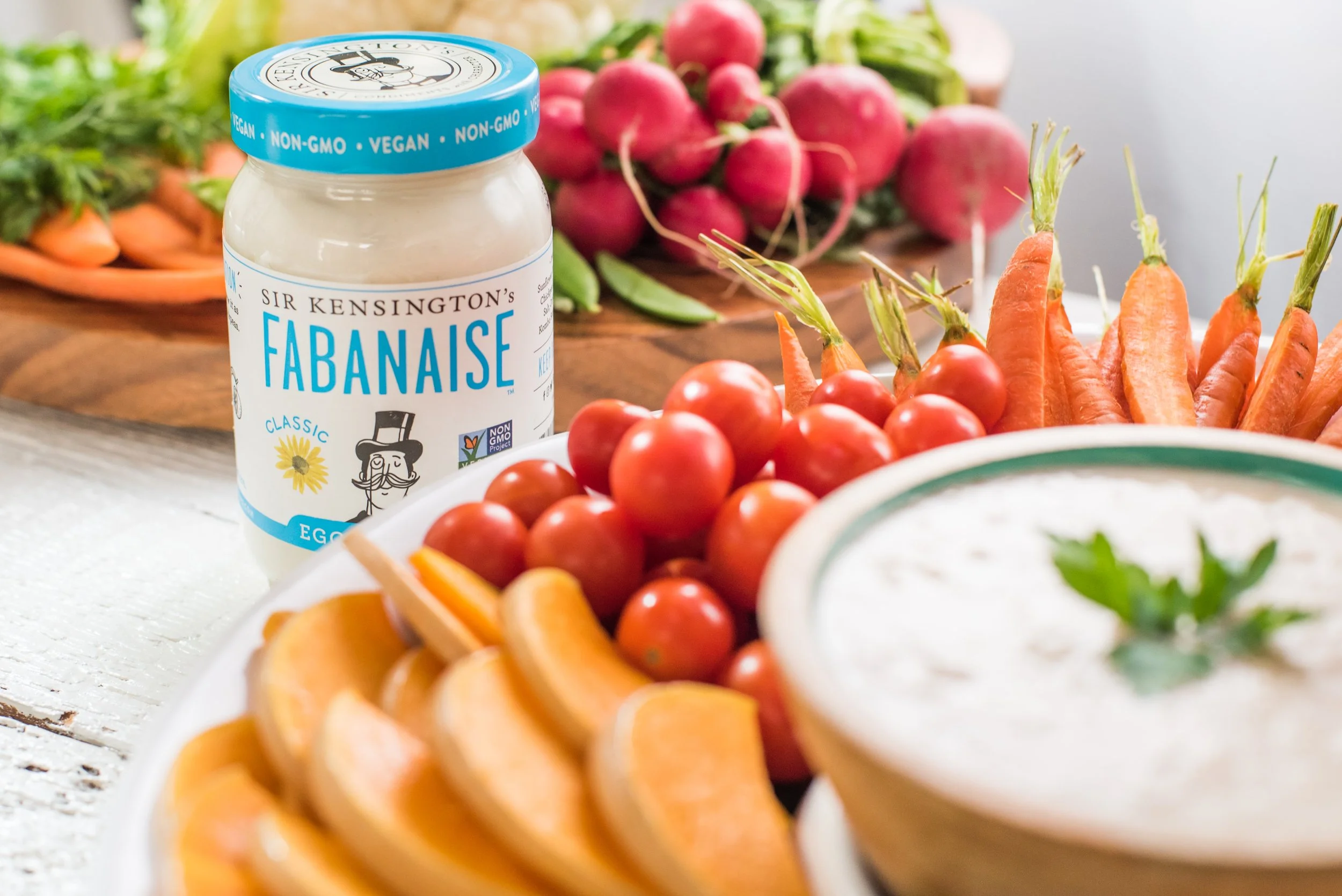 A jar of Sir Kensington's Fabanaise vegan mayonnaise surrounded by grape tomatoes, sliced cantaloupe, baby carrots with greens, and radishes in the background.