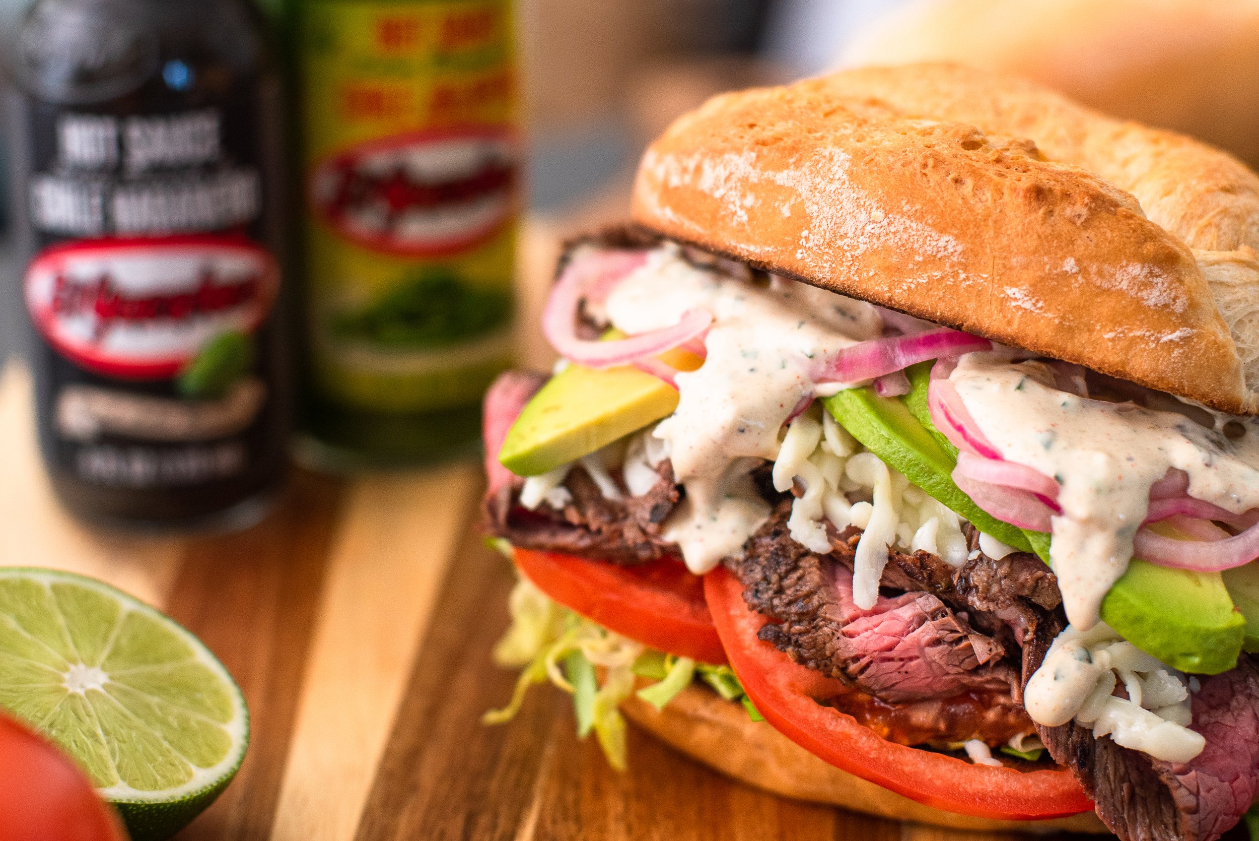 Close-up of a sandwich with beef, tomato, lettuce, onion, avocado, and creamy dressing on a baguette, with lime and canned food in the background.