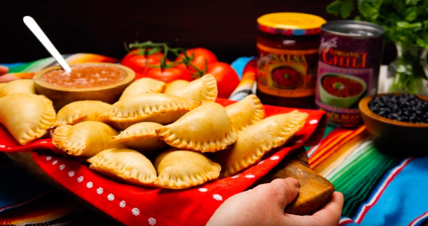 A plate of fried empanadas on a red and white polka dot cloth, with tomatoes, canned chili, jars of salsa, and a bowl of black beans in the background, on a colorful striped tablecloth.
