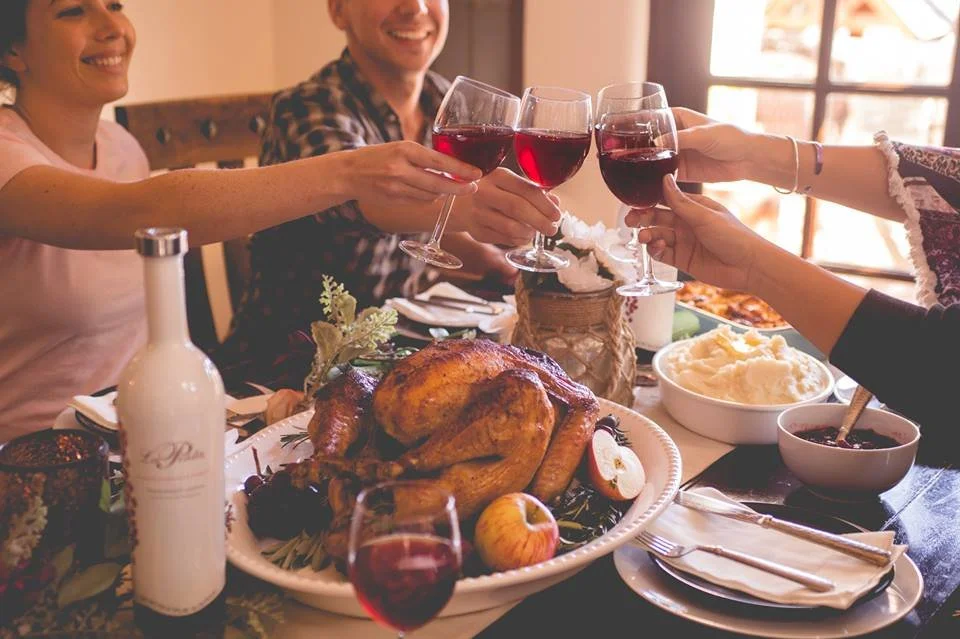 People raising glasses of red wine in a toast over a dinner table with a roasted turkey, mashed potatoes, and other dishes.