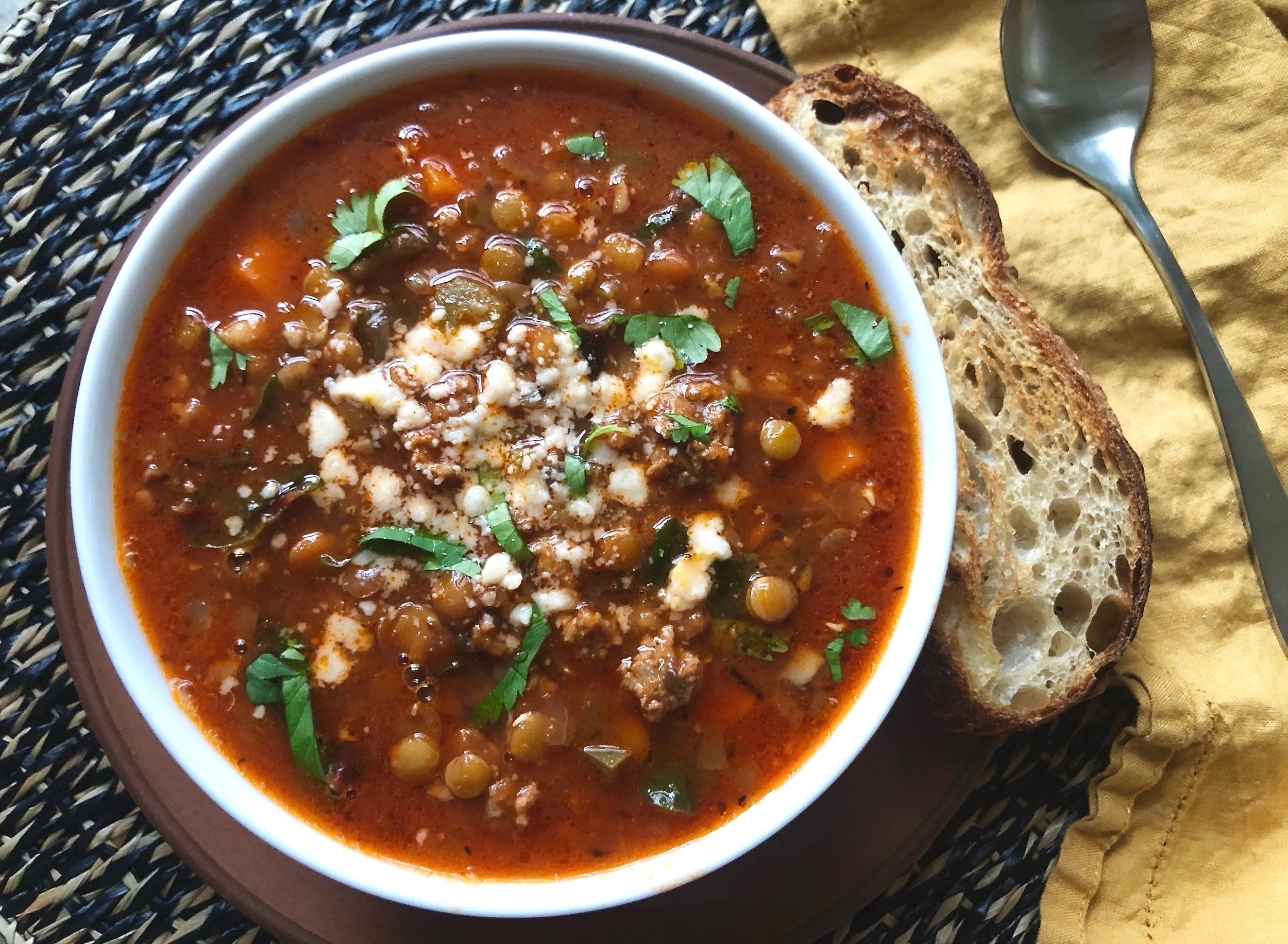 Bowl of lentil soup garnished with herbs, served with slices of toasted bread and a spoon on a yellow napkin.