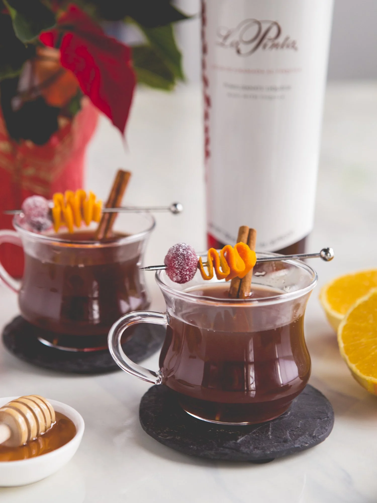 Two glass cups of hot mulled wine garnished with cinnamon sticks, candy, and orange slices on black coasters, with honey, lemon, and a bottle in the background.
