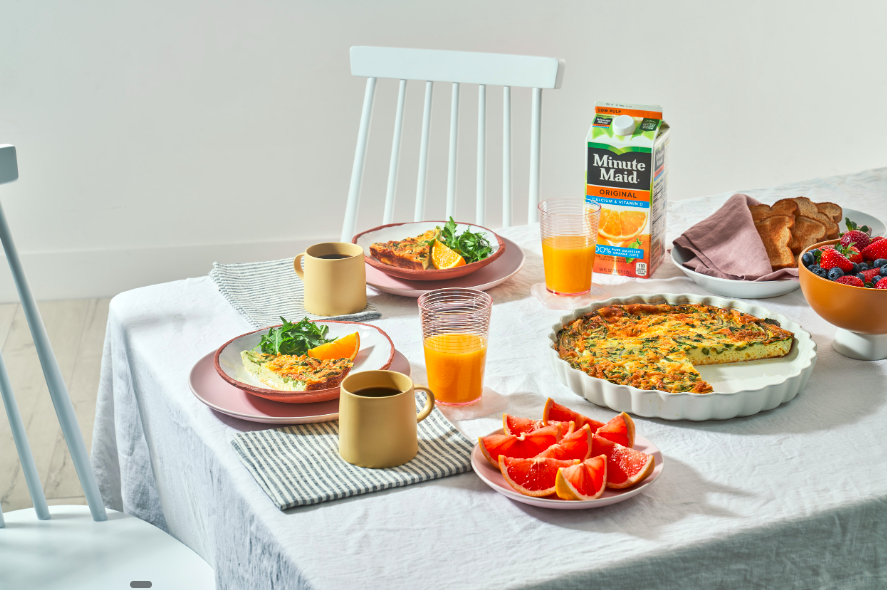 A breakfast table set with two plates of quiche, two cups of coffee, two glasses of orange juice, a plate of grapefruit slices, a carton of Minute Maid orange juice, a bowl of mixed berries, a loaf of bread, and a plate with a slice of quiche on a st