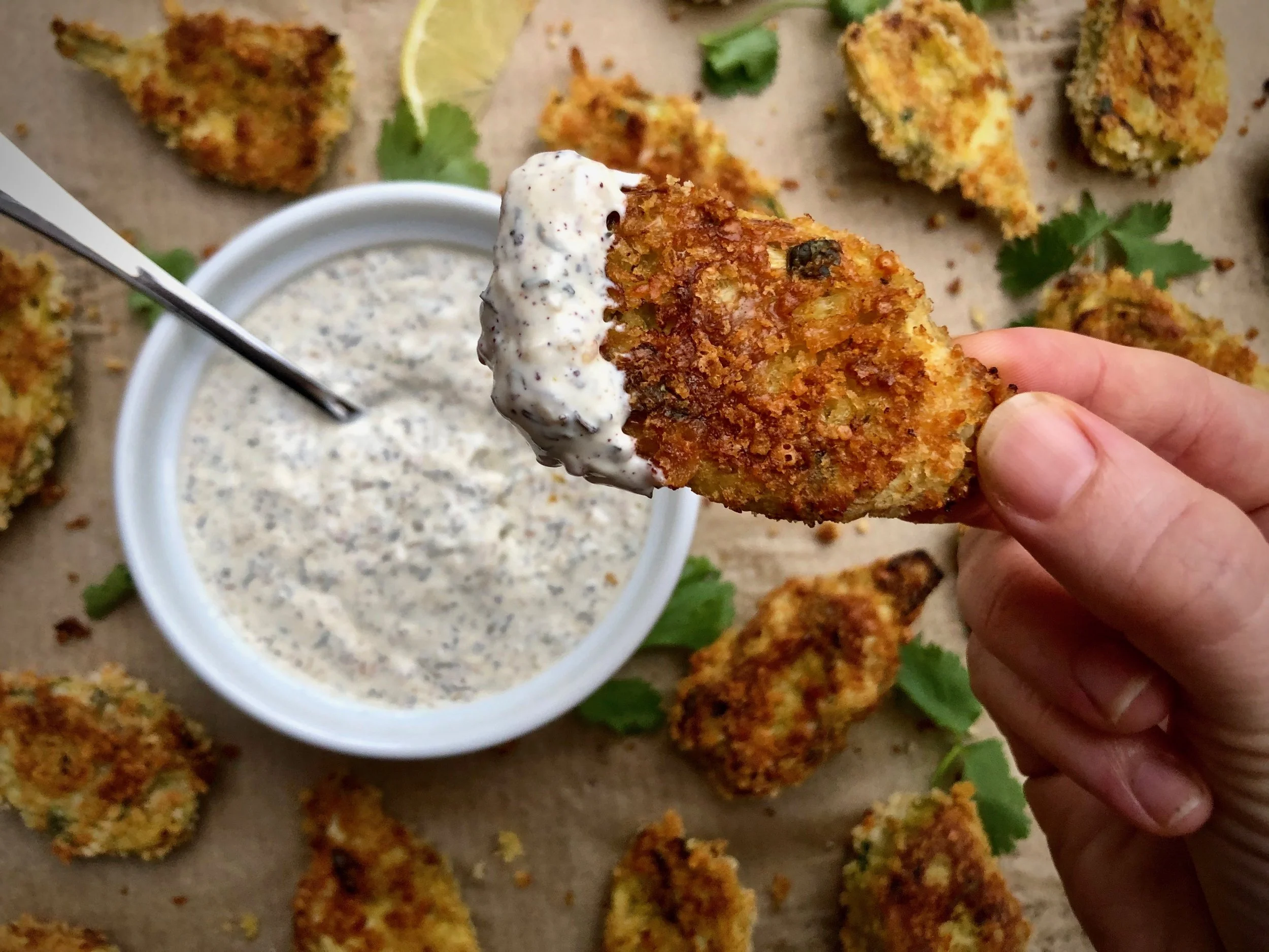 A hand holding a fried chicken wing dipped in ranch dressing, with more fried chicken wings and a bowl of ranch on a brown paper surface, garnished with cilantro and a lime wedge.