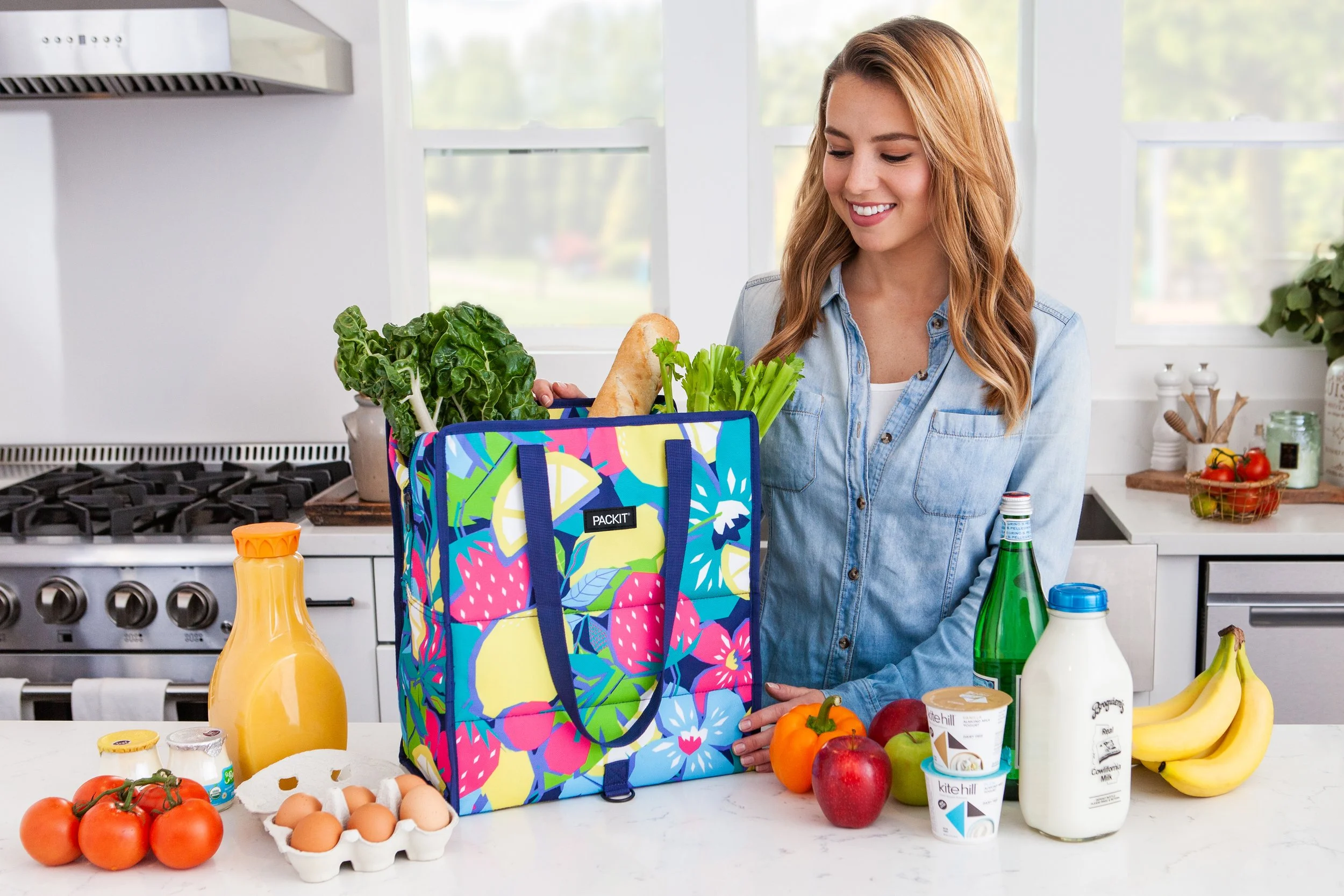 A woman packing a colorful tote bag with groceries on a kitchen countertop, including leafy greens, bread, and other food items, with fresh produce like tomatoes, bananas, apples, orange, and milk nearby.